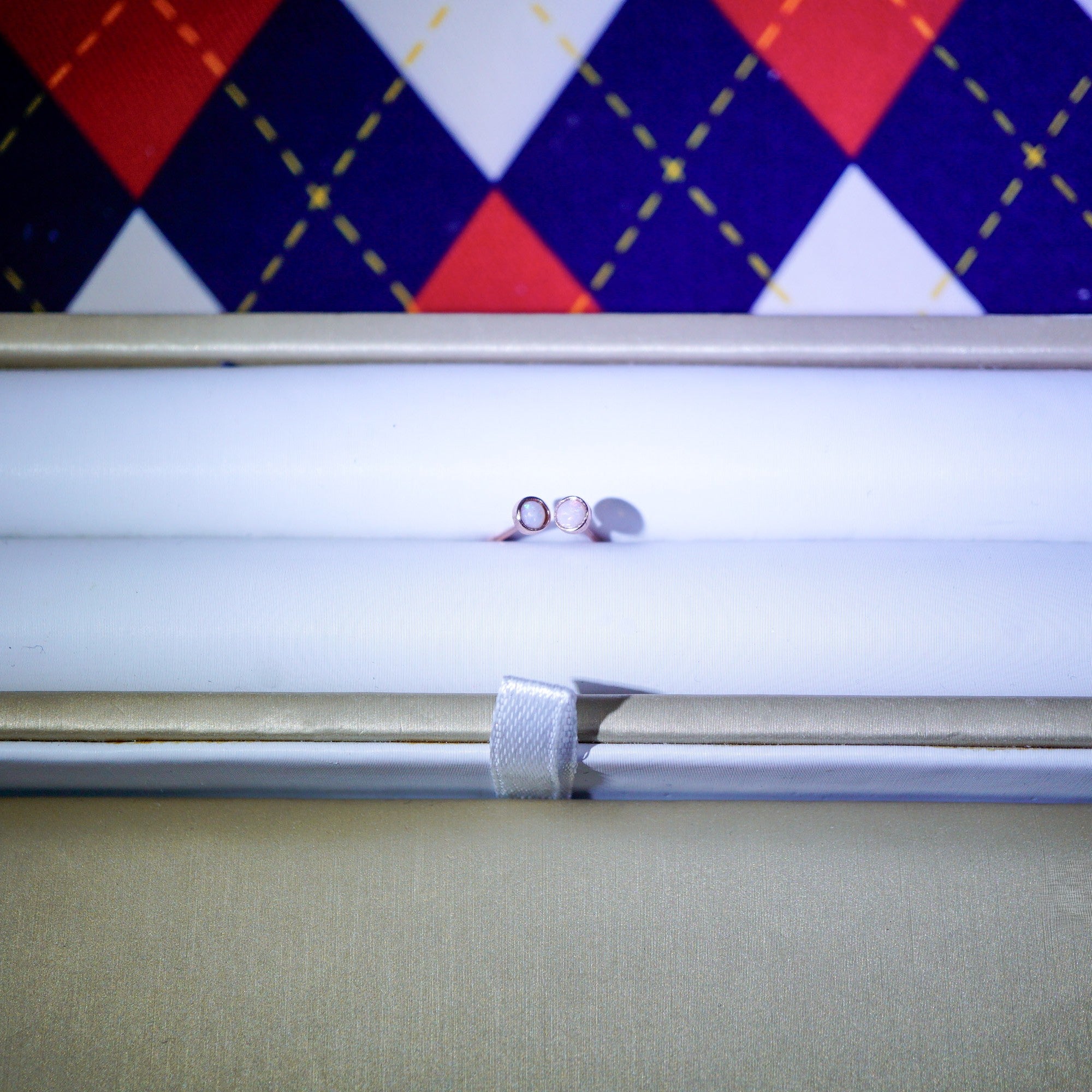 A pair of small silver hoop earrings displayed horizontally inside an open white jewelry box, with a colorful argyle-patterned fabric background visible above.