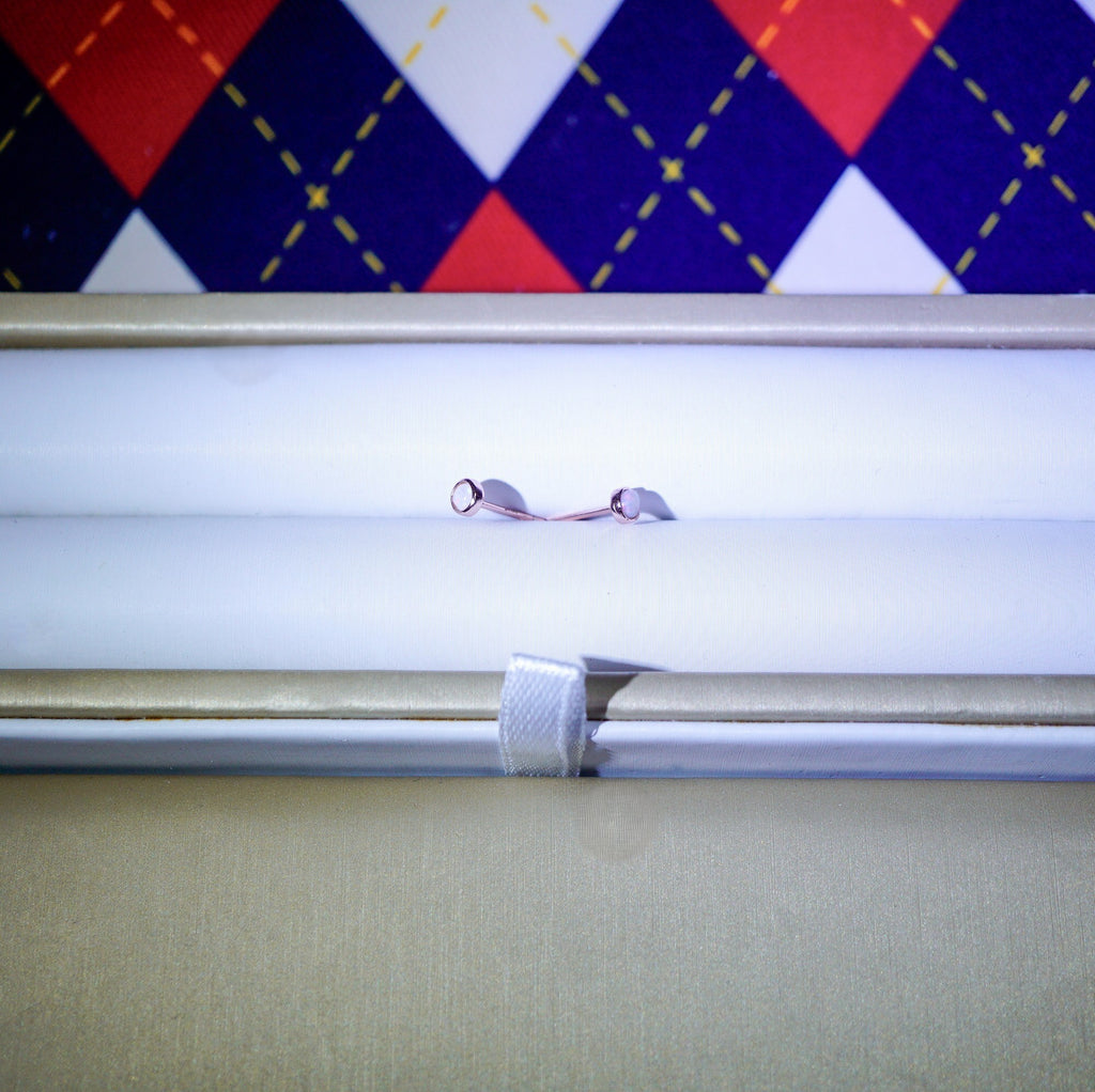 A close-up of a small metallic jewelry piece, possibly a ring or chain with two small silver-toned loops, resting on a white surface inside a beige box. A red, navy, and white argyle patterned fabric is visible in the background.