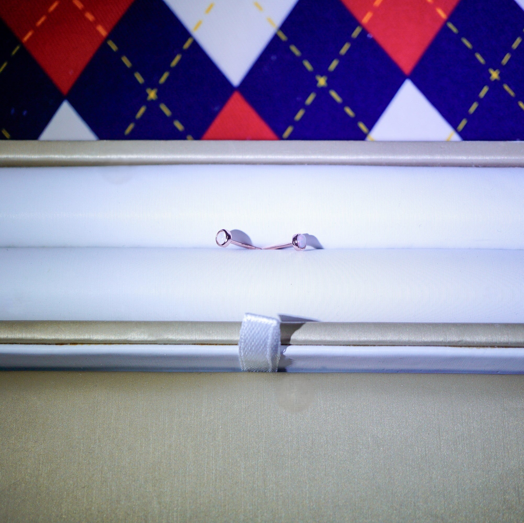 A close-up of a small metallic jewelry piece, possibly a ring or chain with two small silver-toned loops, resting on a white surface inside a beige box. A red, navy, and white argyle patterned fabric is visible in the background.