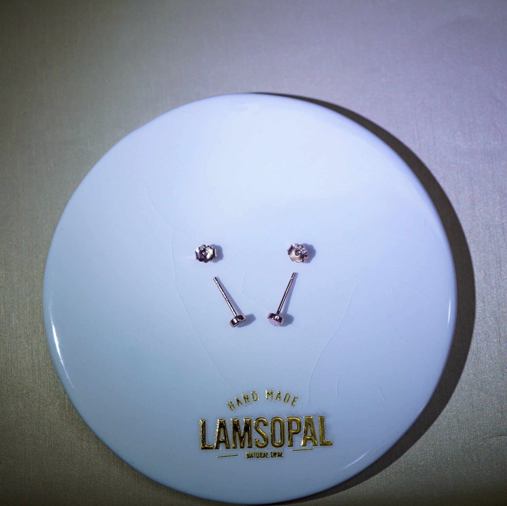 Two small silver-toned stud earrings displayed on a round white backing card with the brand name LAMSOPAL printed in gold at the bottom.