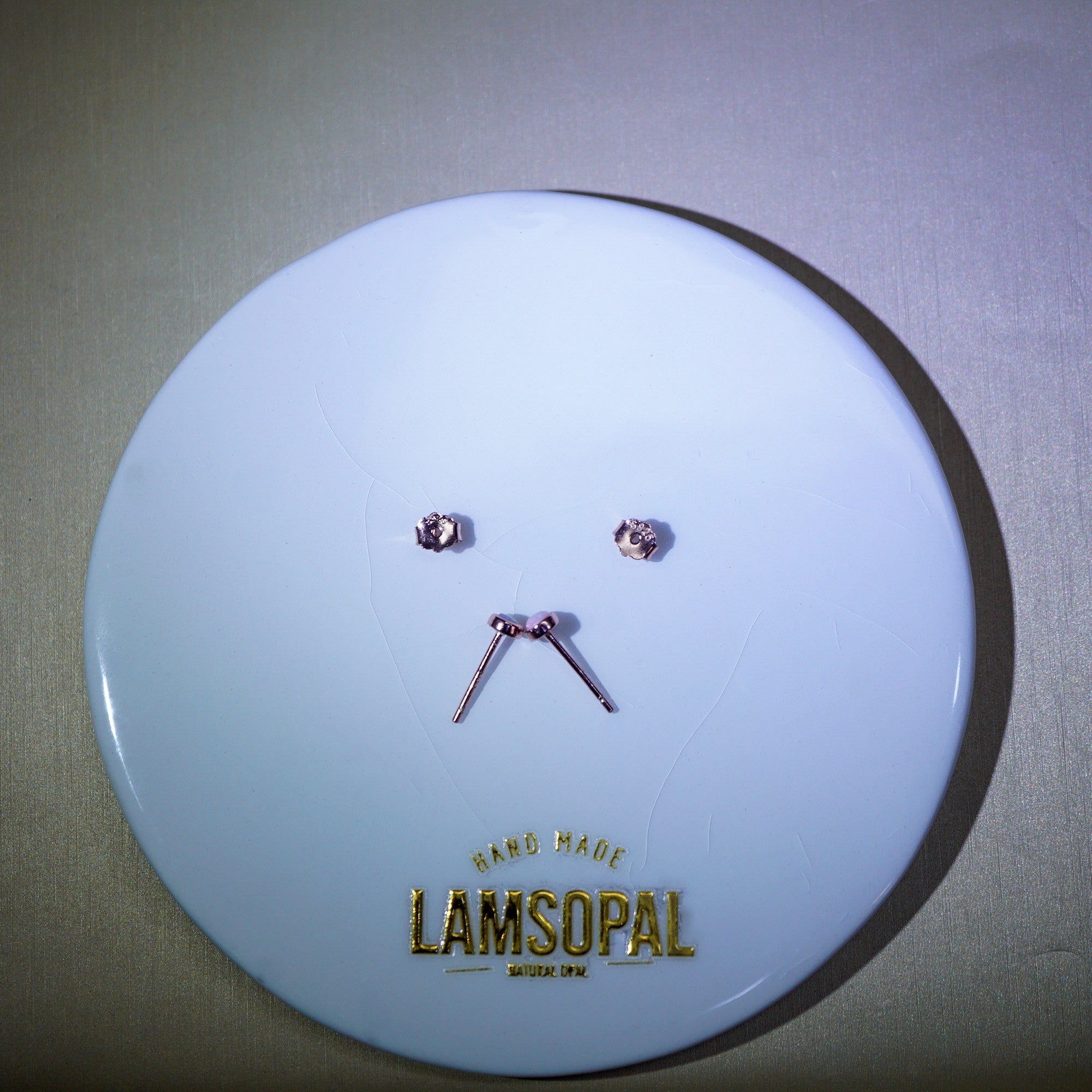 A pair of small rose gold stud earrings and matching nose studs displayed on a round white Lamsopal brand card against a neutral gray background.
