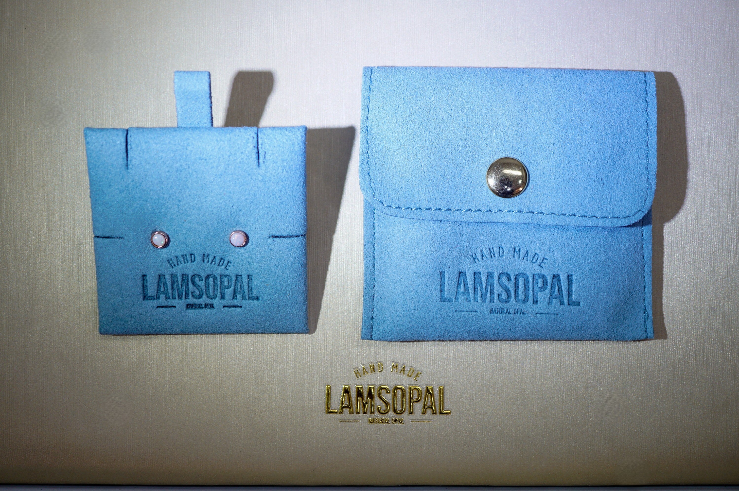 A pair of light blue handmade leather card holders or wallet-like accessories with the brand name LAMSOPAL embossed on them, displayed on a beige background.
