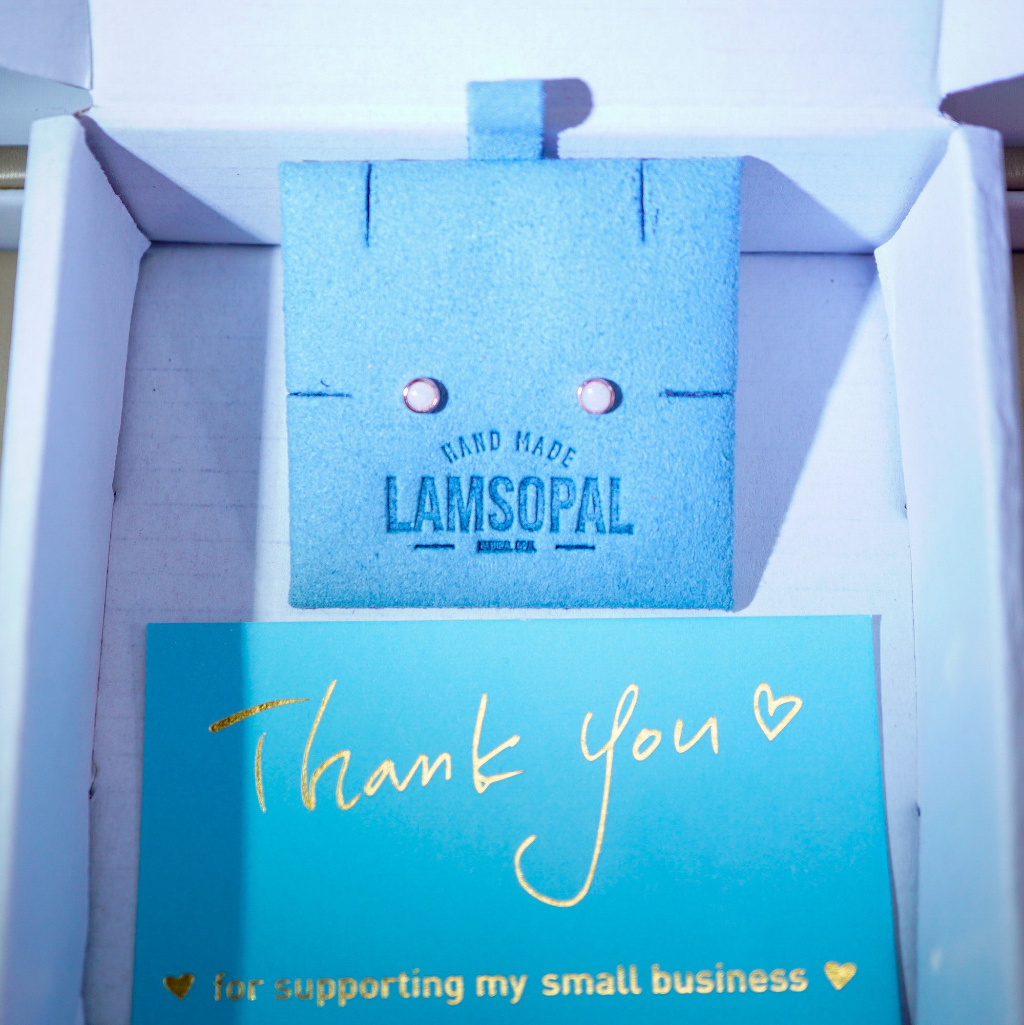 A small blue textured jewelry box or display insert with the words 'HAND MADE LAMSOPAL' printed on it, placed inside a white box next to a turquoise thank-you card with gold script reading 'Thank you' and 'i'm supporting my small business'.