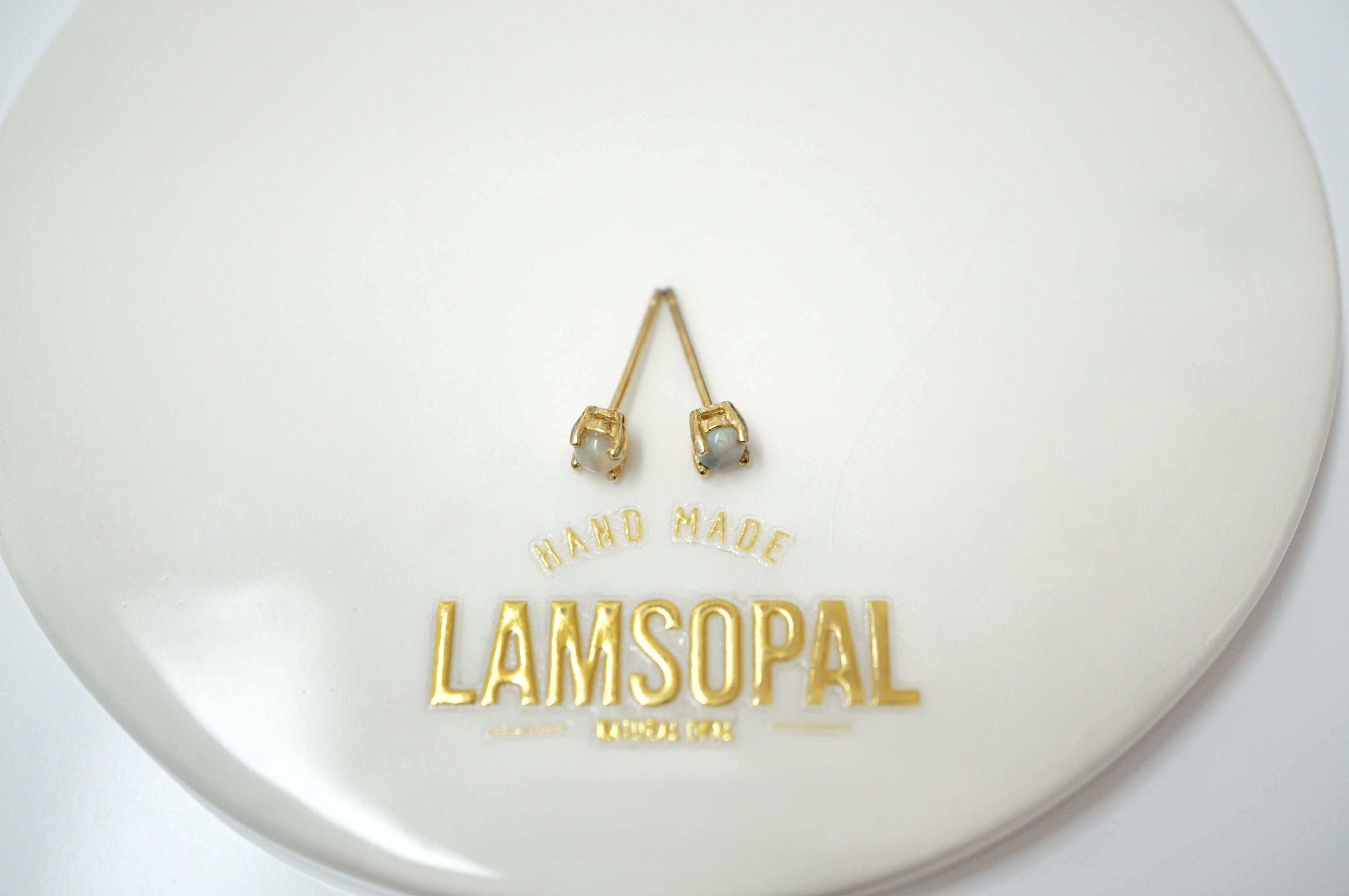A pair of gold-colored stud earrings with small clear stones, displayed on a white circular surface with gold foil branding that reads 'HAND MADE LAMSOPAL'.