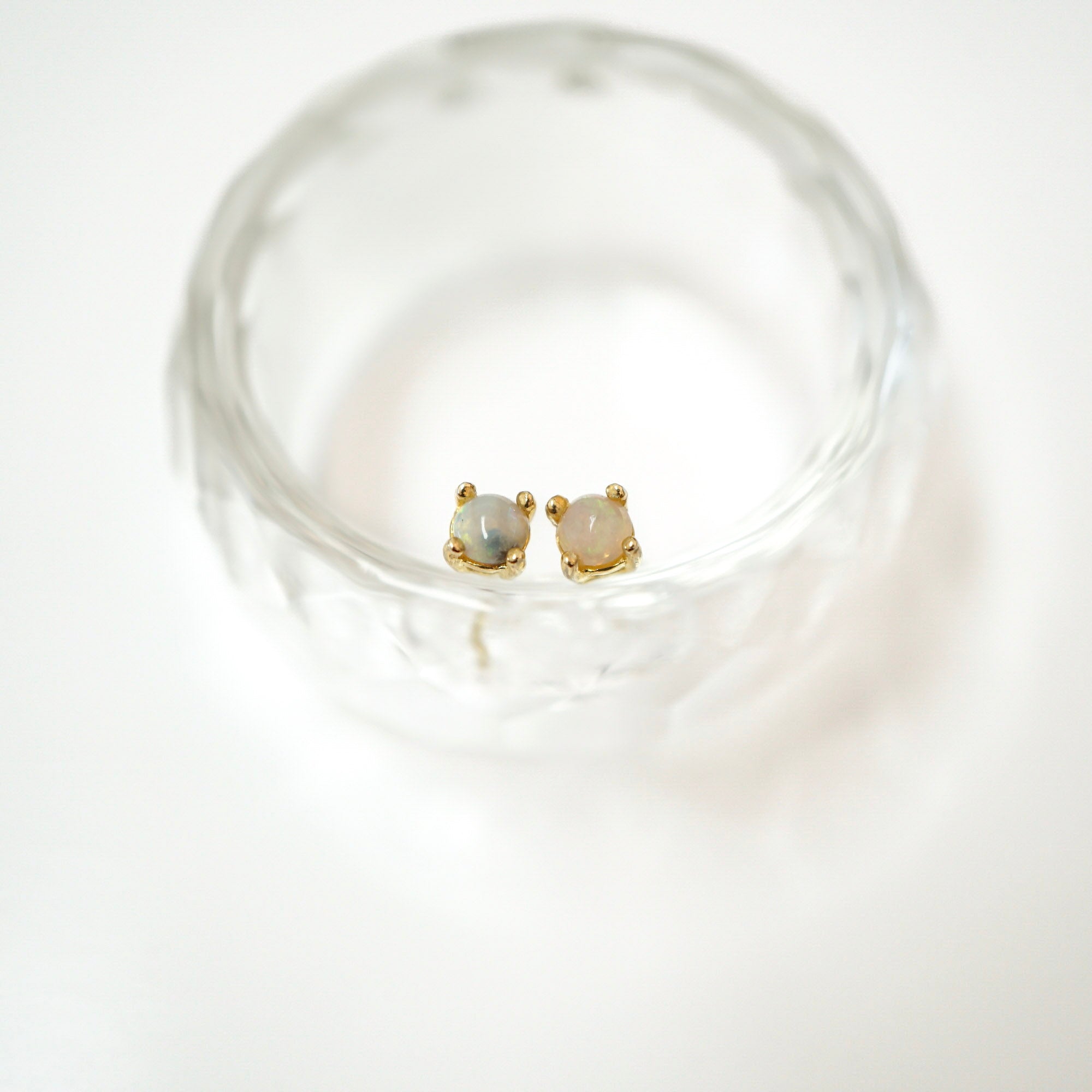 A pair of small gold stud earrings with oval milky white stones, displayed inside a clear textured glass bowl on a white background.