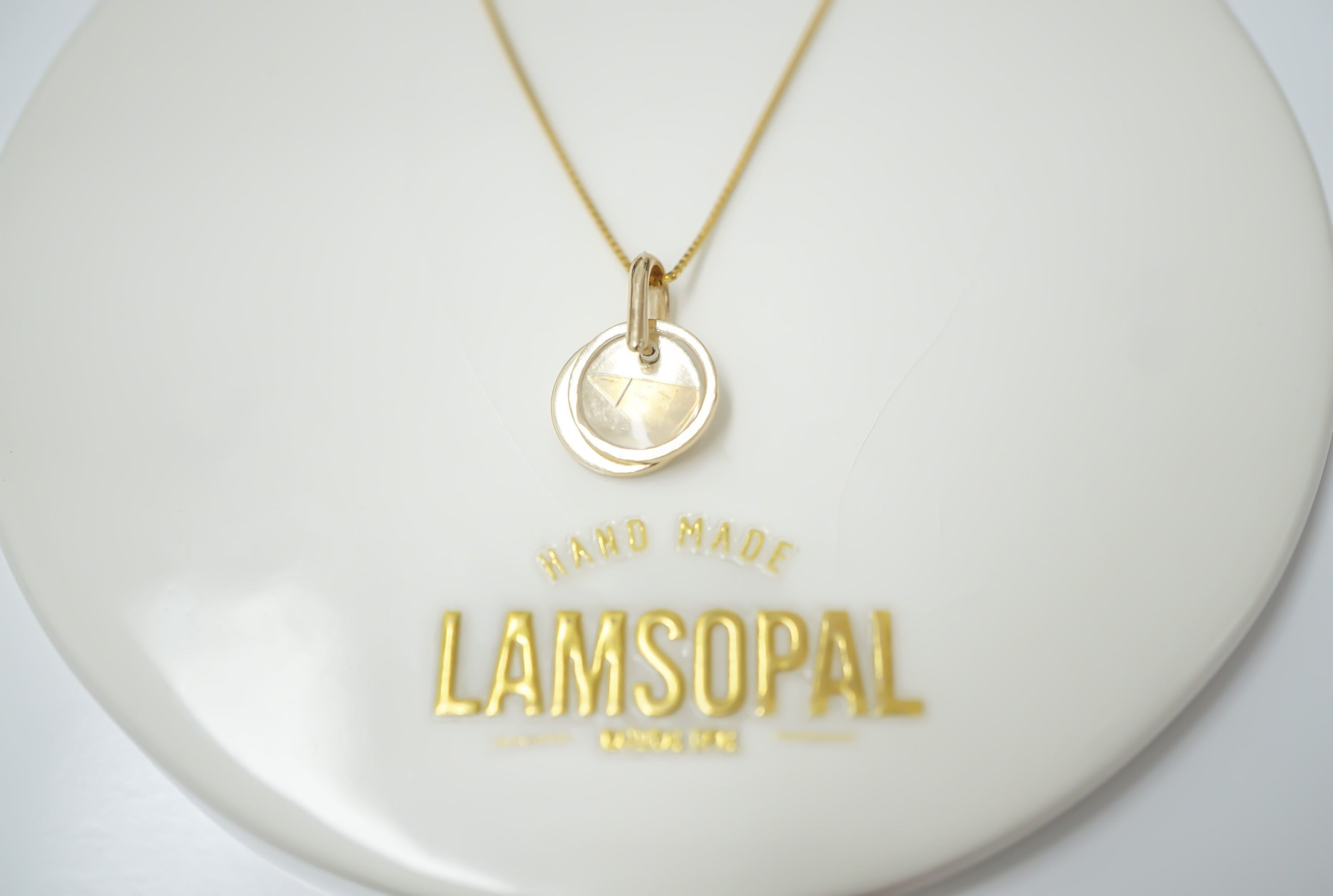 A gold pendant necklace with a circular disc charm featuring a star design, displayed on a white circular stand with the brand name 'LAMSOPAL' printed in gold.