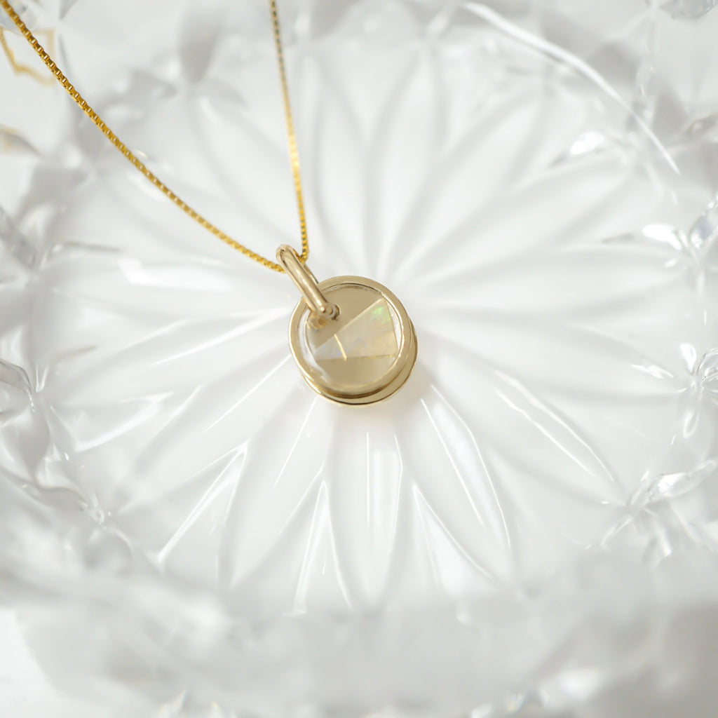 A gold necklace with a round bezel-set iridescent stone pendant, displayed on a textured white glass dish.