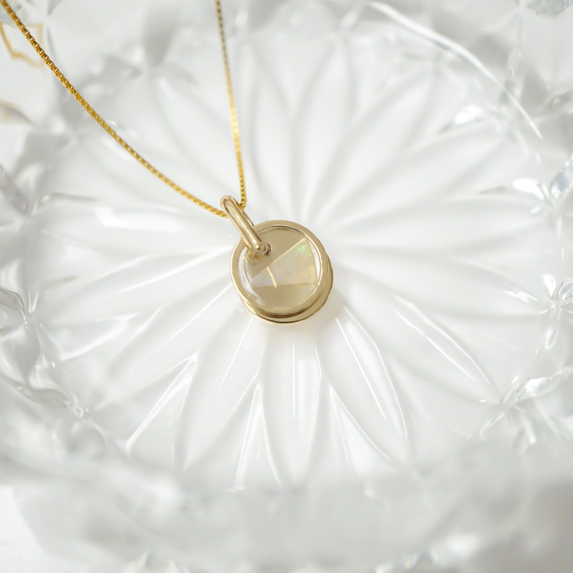 A gold necklace with a round bezel-set iridescent stone pendant, displayed on a textured white glass dish.