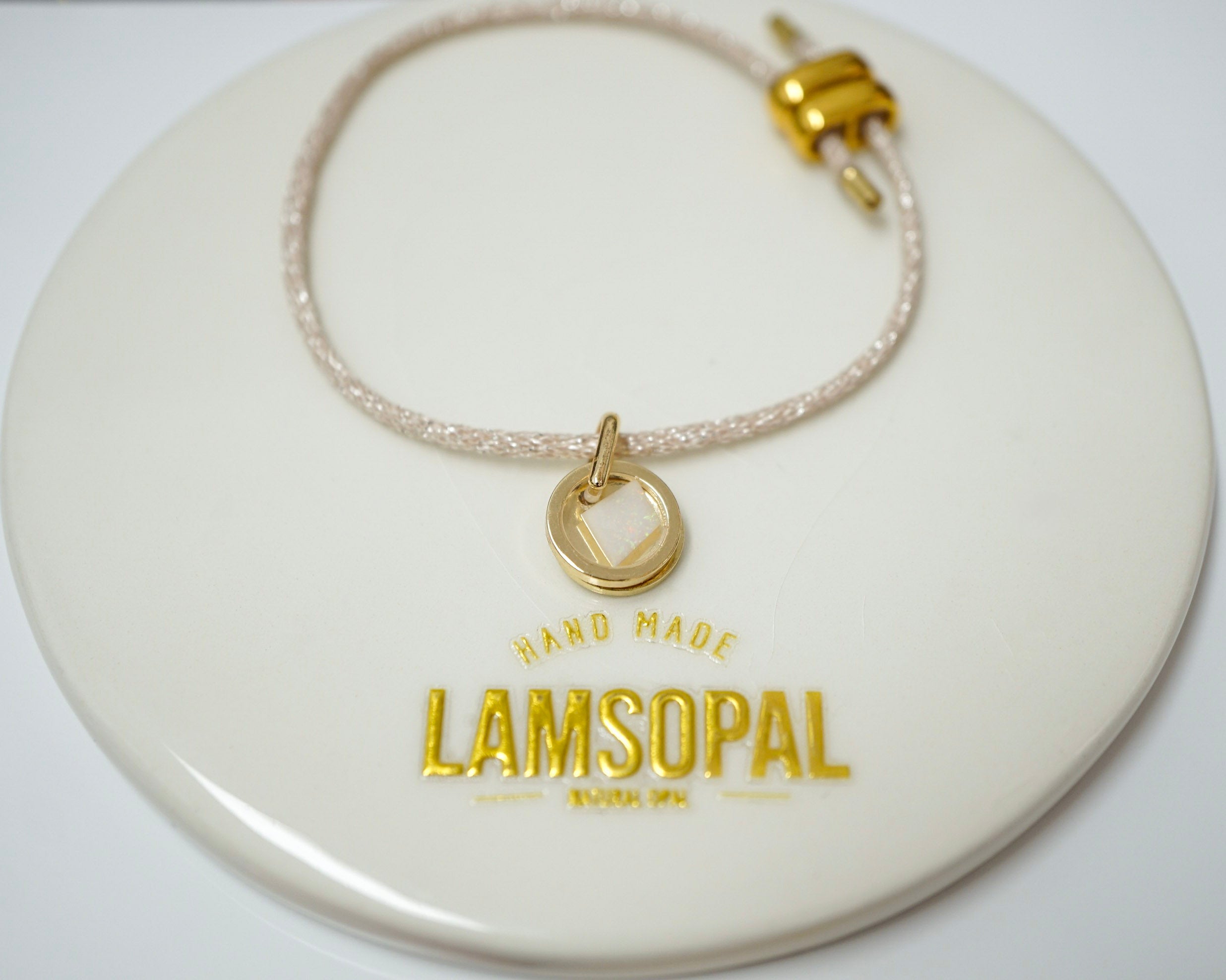 A delicate beaded bracelet with a gold heart-shaped charm and clasp, displayed on a round cream-colored Lamsopal brand card.