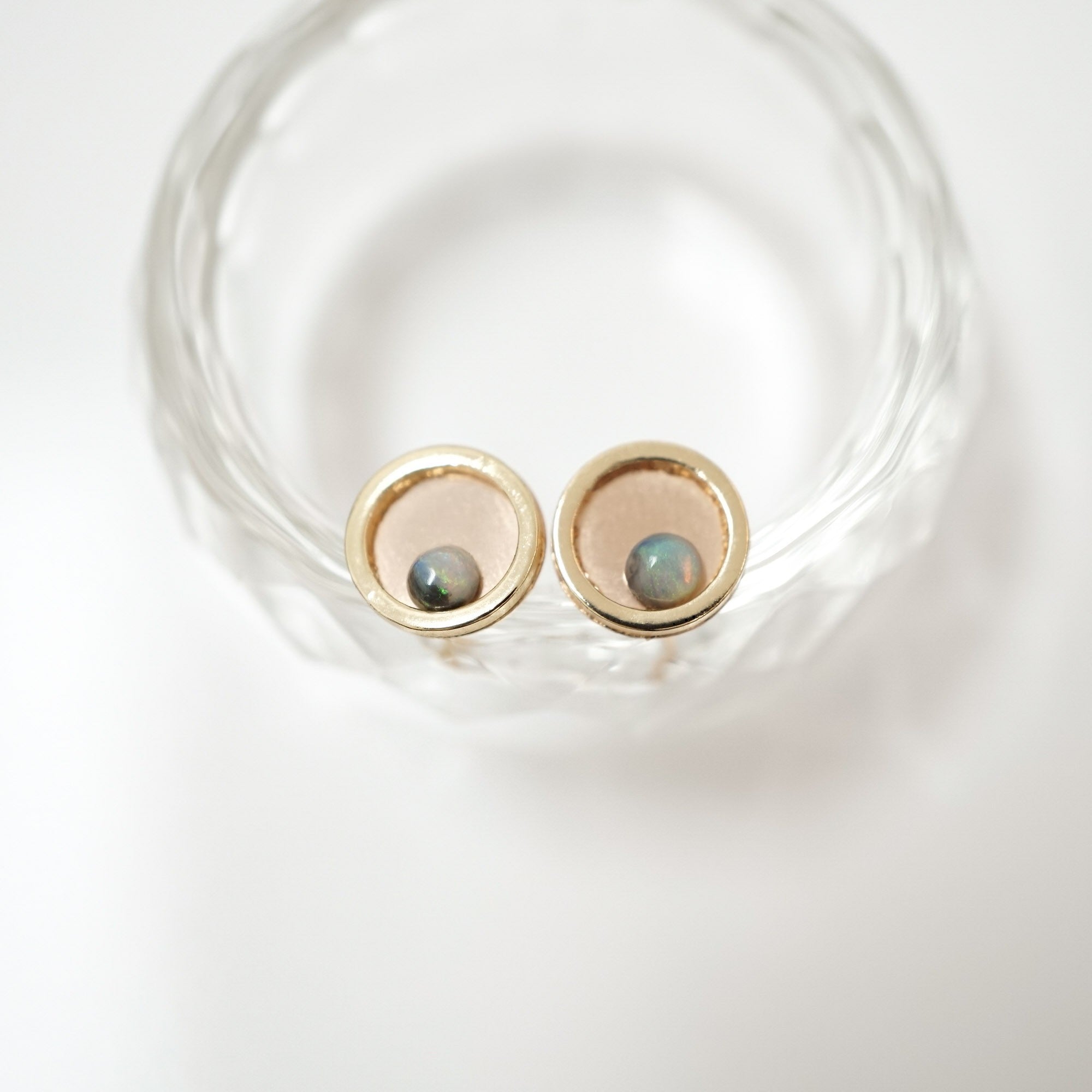 A pair of gold-toned stud earrings with oval bezel settings holding iridescent blue-green cabochon stones, displayed inside a clear textured glass bowl on a white background.