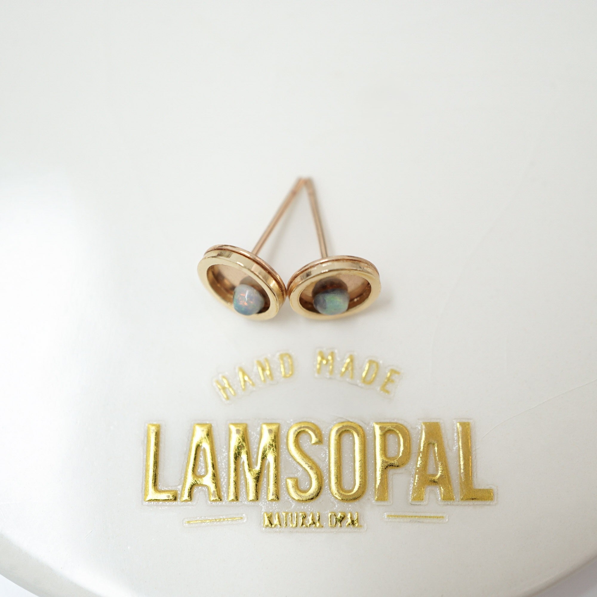 A pair of gold-toned oval stud earrings with small natural opal inlays, displayed on a white surface above gold foil text reading 'HAND MADE LAMSOPAL'.