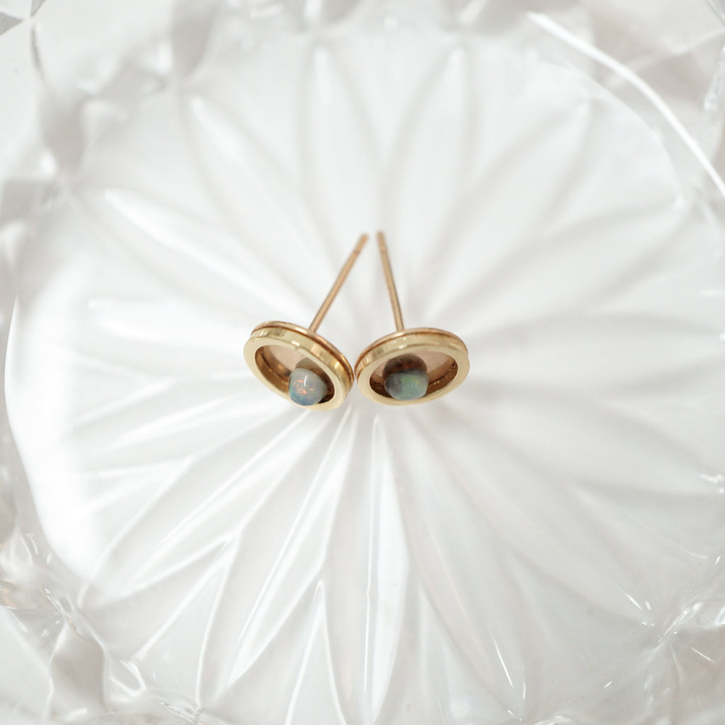 A pair of gold stud earrings with oval bezel settings holding small round iridescent blue-green cabochon stones, displayed on a textured white glass dish.