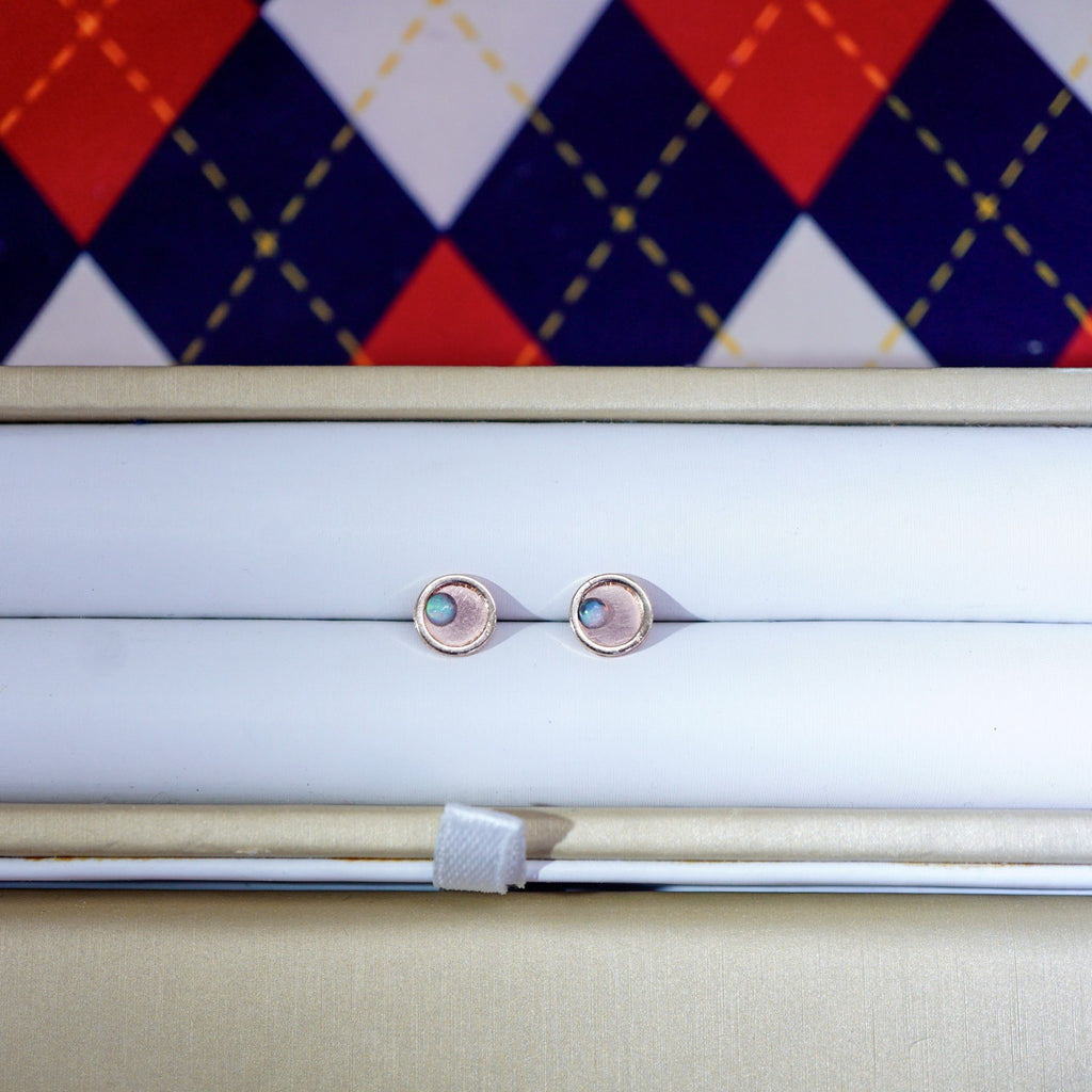 A pair of small oval stud earrings with iridescent inlay centers and rose-gold-toned edges, displayed on a white cushion inside a beige jewelry box. A red, navy, and white argyle pattern is visible in the background.