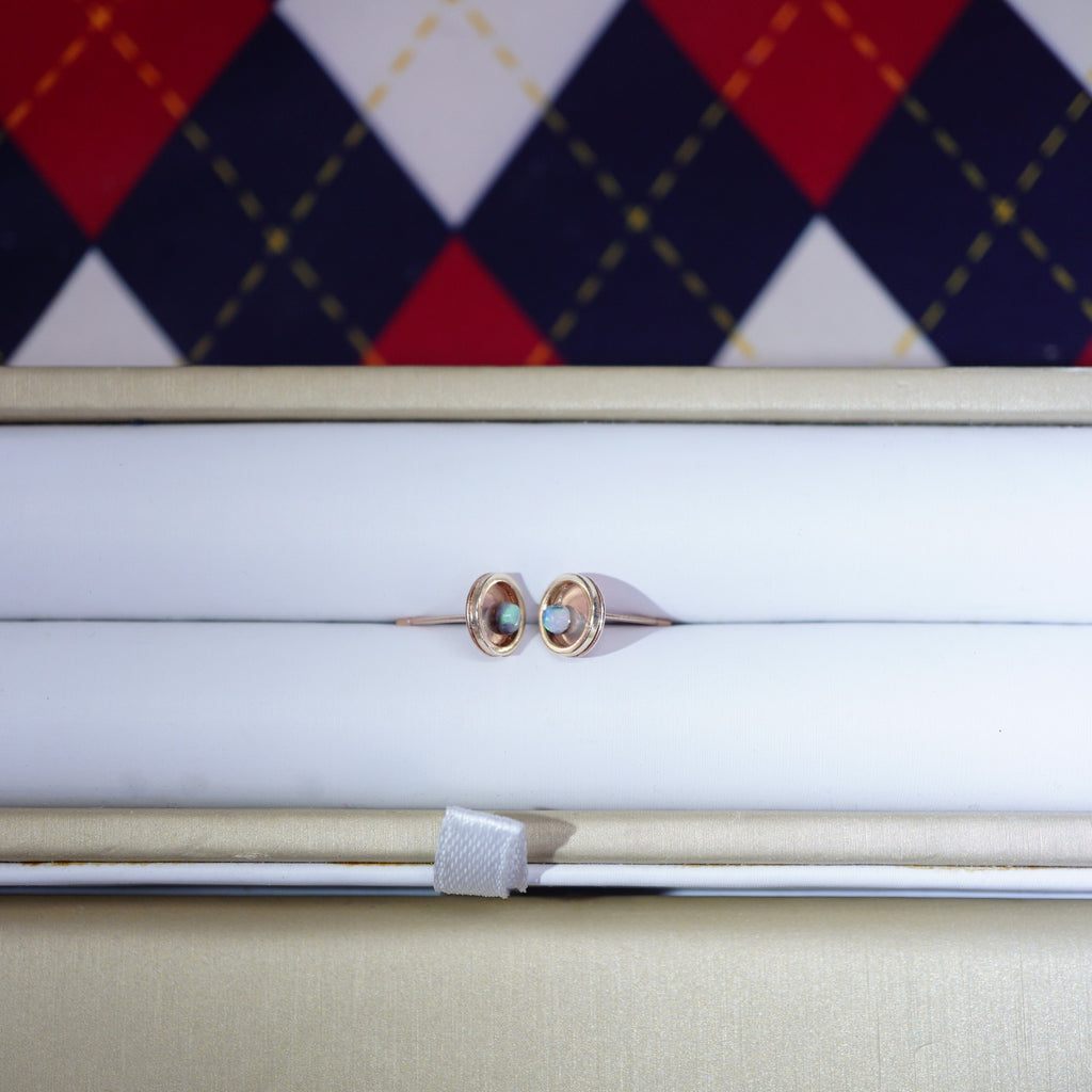 A pair of rose gold hoop earrings with small oval-cut light blue gemstones, presented in a white jewelry box with a beige outer lid. A red, navy, and white argyle patterned fabric is visible in the background.