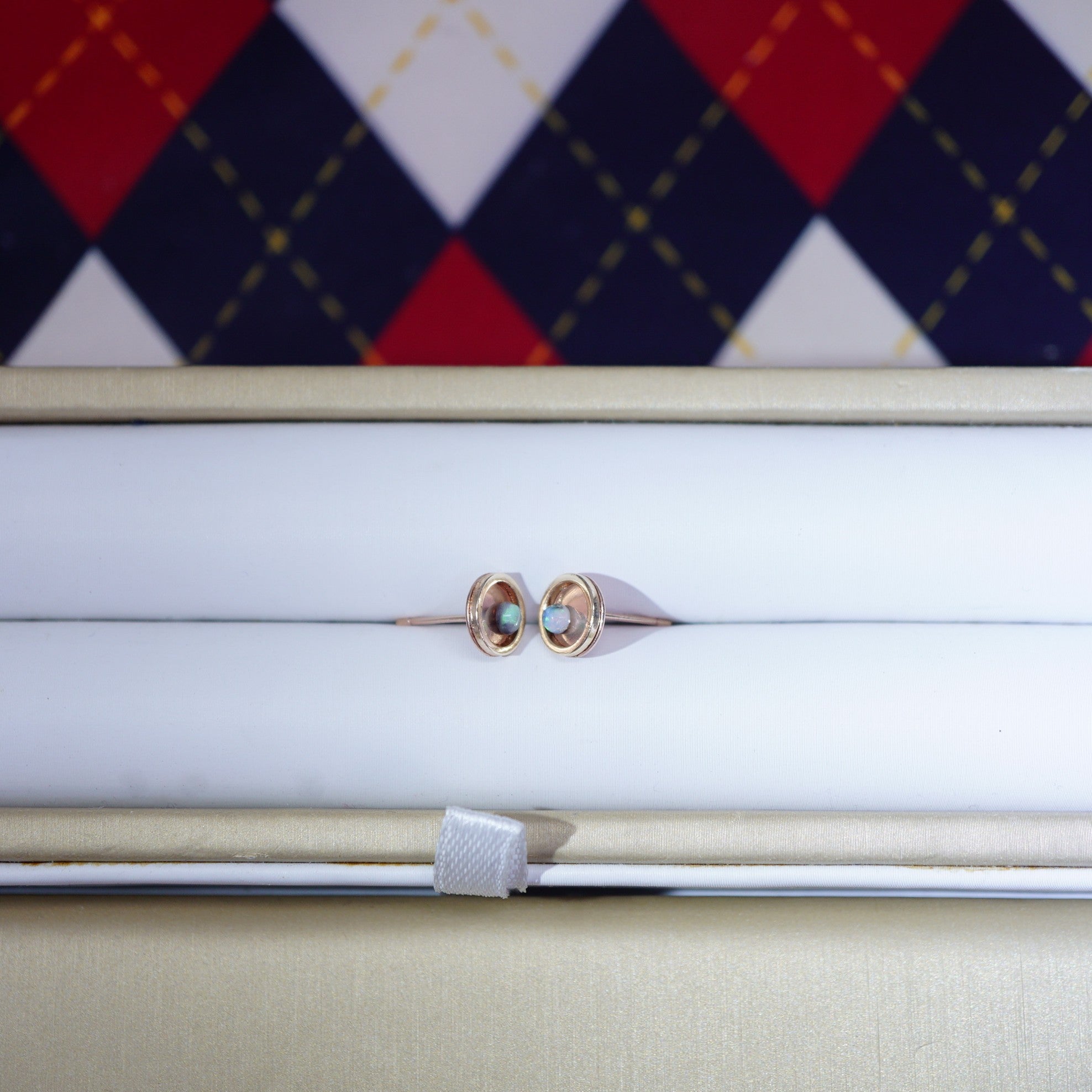 A pair of rose gold hoop earrings with small oval-cut light blue gemstones, presented in a white jewelry box with a beige outer lid. A red, navy, and white argyle patterned fabric is visible in the background.