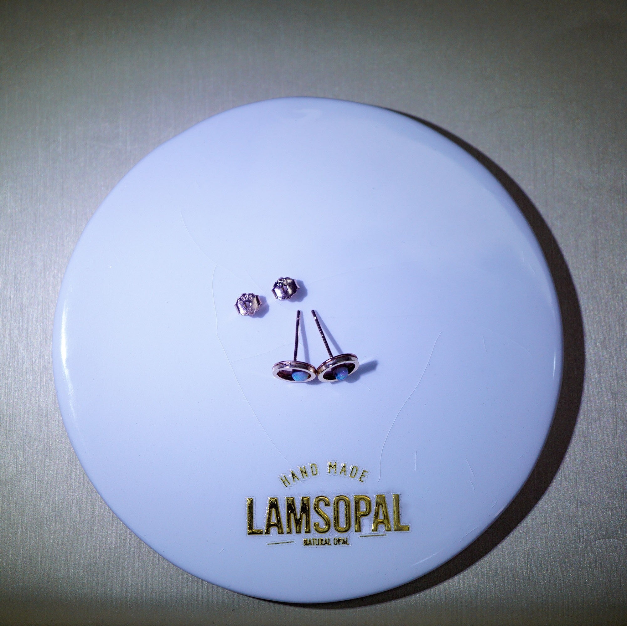 A pair of rose gold stud earrings with clear round-cut stones and matching backings, displayed on a white circular branded card that reads 'HAND MADE LAMSOPAL'.