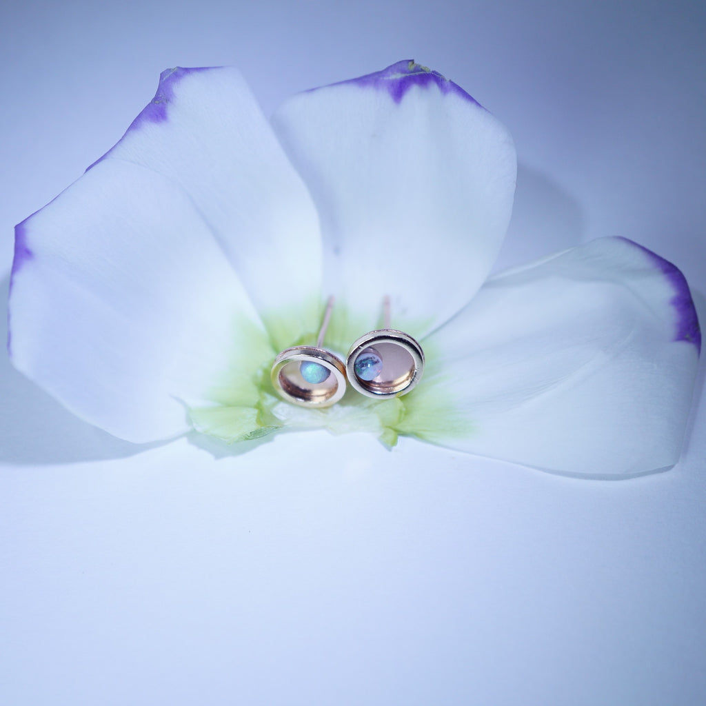 A pair of gold-toned oval stud earrings with iridescent blue-green opal-like centers, displayed on the petals of a white orchid flower against a pale blue background.