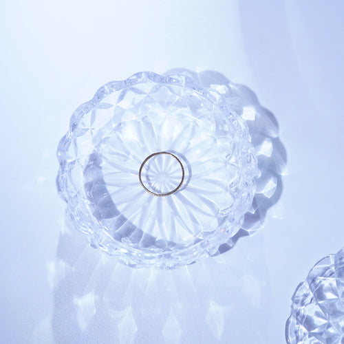 A clear faceted glass bowl with a scalloped rim and a small metallic ring in the center, photographed on a pale blue-white background with soft reflections.