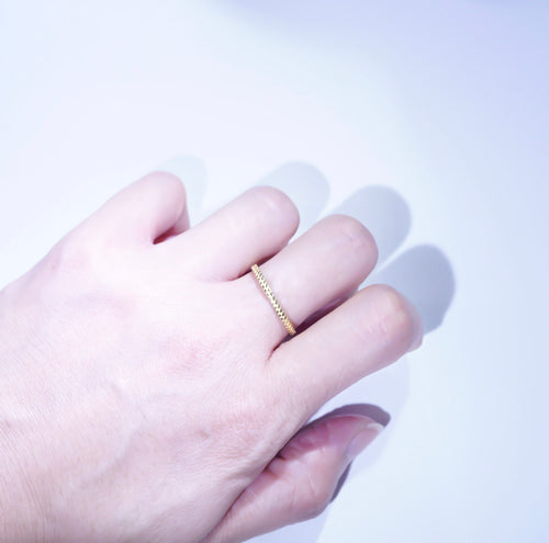A close-up of a hand wearing a thin textured gold band ring on one finger, set against a bright white background with soft shadows.