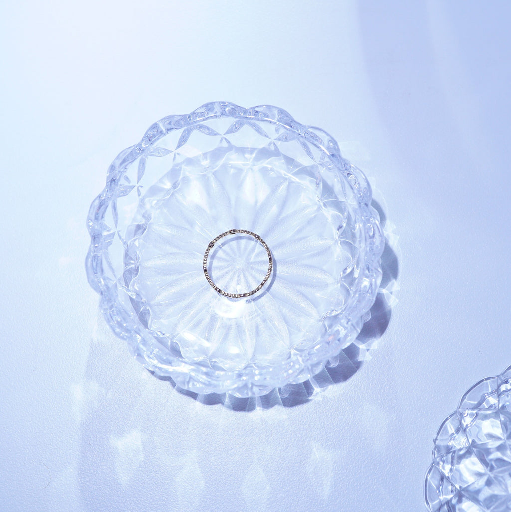A delicate silver ring with small clear stones, displayed in the center of a clear, scalloped glass dish on a light blue-white surface.