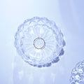 A delicate silver ring with small clear stones, displayed in the center of a clear, scalloped glass dish on a light blue-white surface.