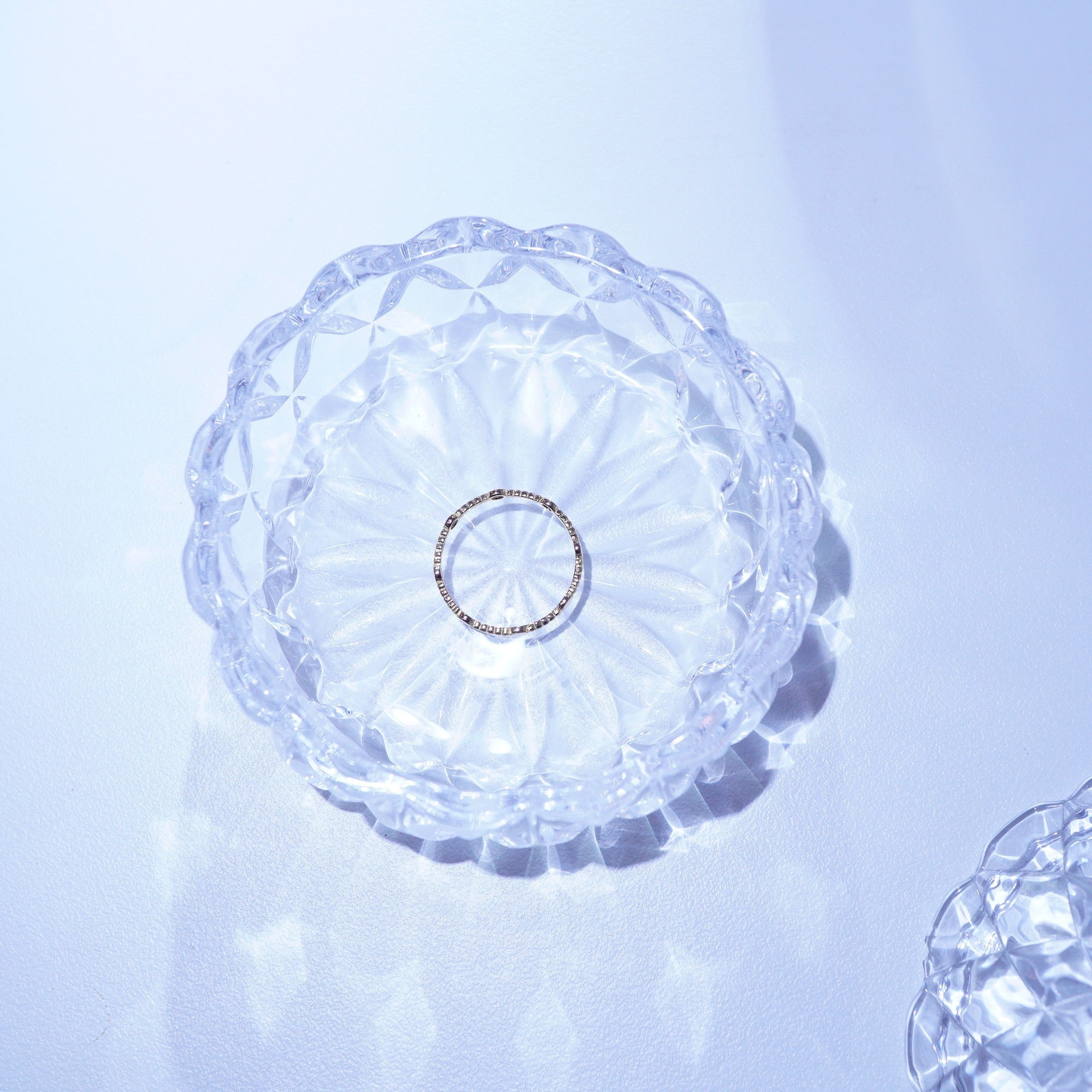 A delicate silver ring with small clear stones, displayed in the center of a clear, scalloped glass dish on a light blue-white surface.