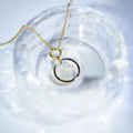A gold chain necklace with a double-ring pendant, one of which is a clear faceted gemstone, displayed inside a textured clear glass bowl.