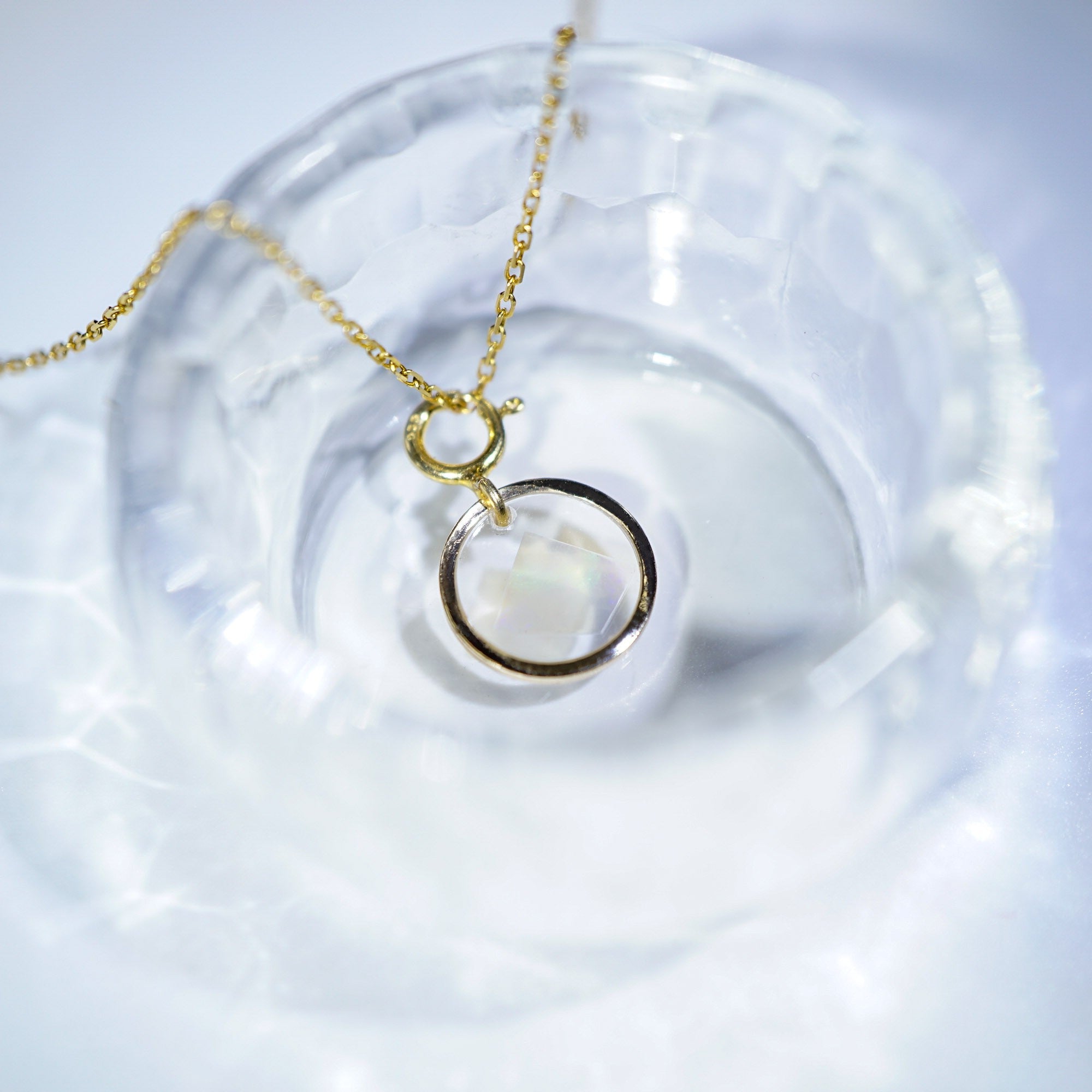 A gold chain necklace with a double-ring pendant, one of which is a clear faceted gemstone, displayed inside a textured clear glass bowl.