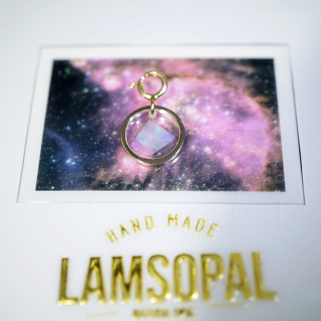 A gold-toned circular pendant charm with a faceted iridescent stone, displayed on a printed card featuring a galaxy background. The card includes the text 'HAND MADE' and 'LAMSOPAL'.