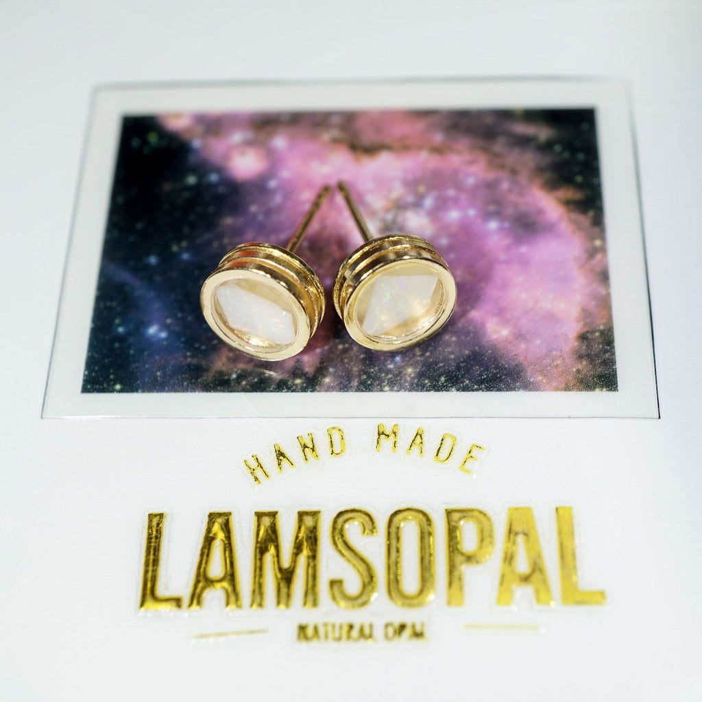 A pair of gold stud earrings with oval milky white inlays, displayed on a card featuring a purple and blue galaxy background. The card is labeled 'HAND MADE LAMSOPAL NATURAL OPAL'.