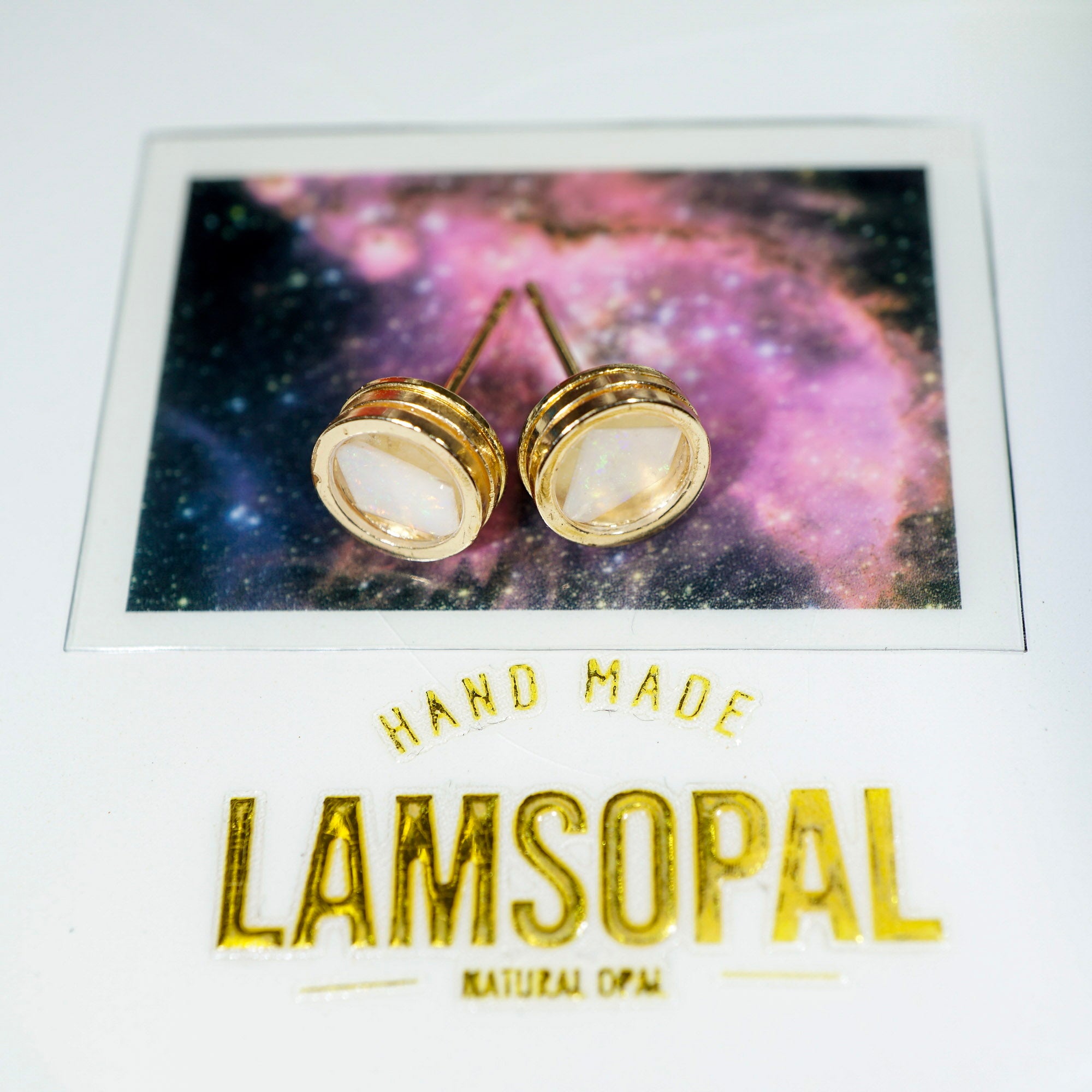 A pair of gold stud earrings with oval milky white inlays, displayed on a card featuring a purple and blue galaxy background. The card is labeled 'HAND MADE LAMSOPAL NATURAL OPAL'.
