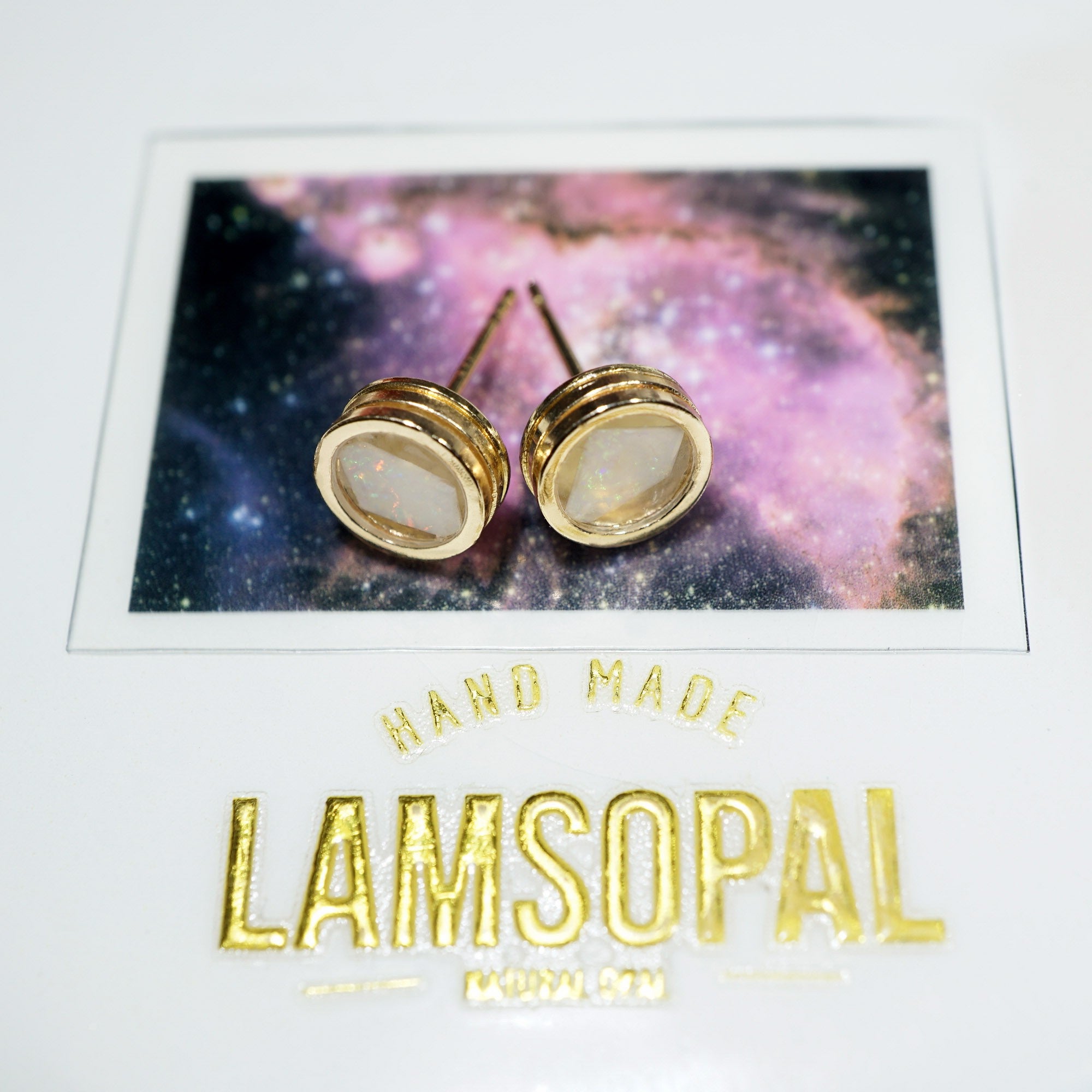 A pair of gold post stud earrings with oval milky white inlays, displayed on a branded white backing card featuring a galaxy print and the brand name Lamsopal.
