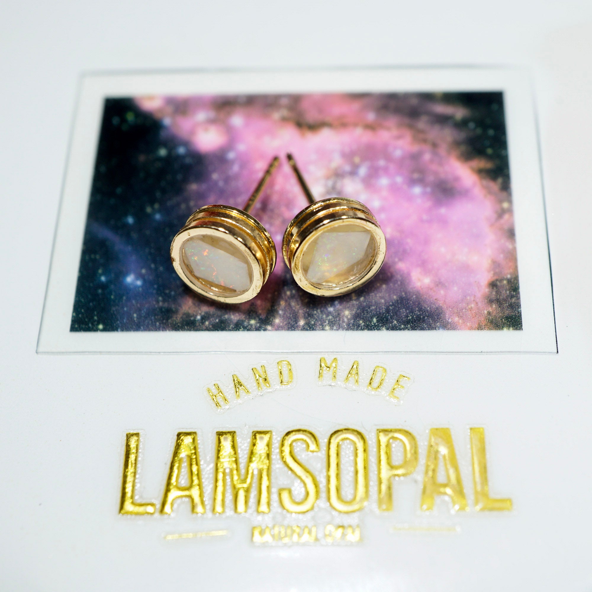 A pair of gold post stud earrings with oval milky white inlays, displayed on a branded white backing card featuring a galaxy print and the brand name Lamsopal.