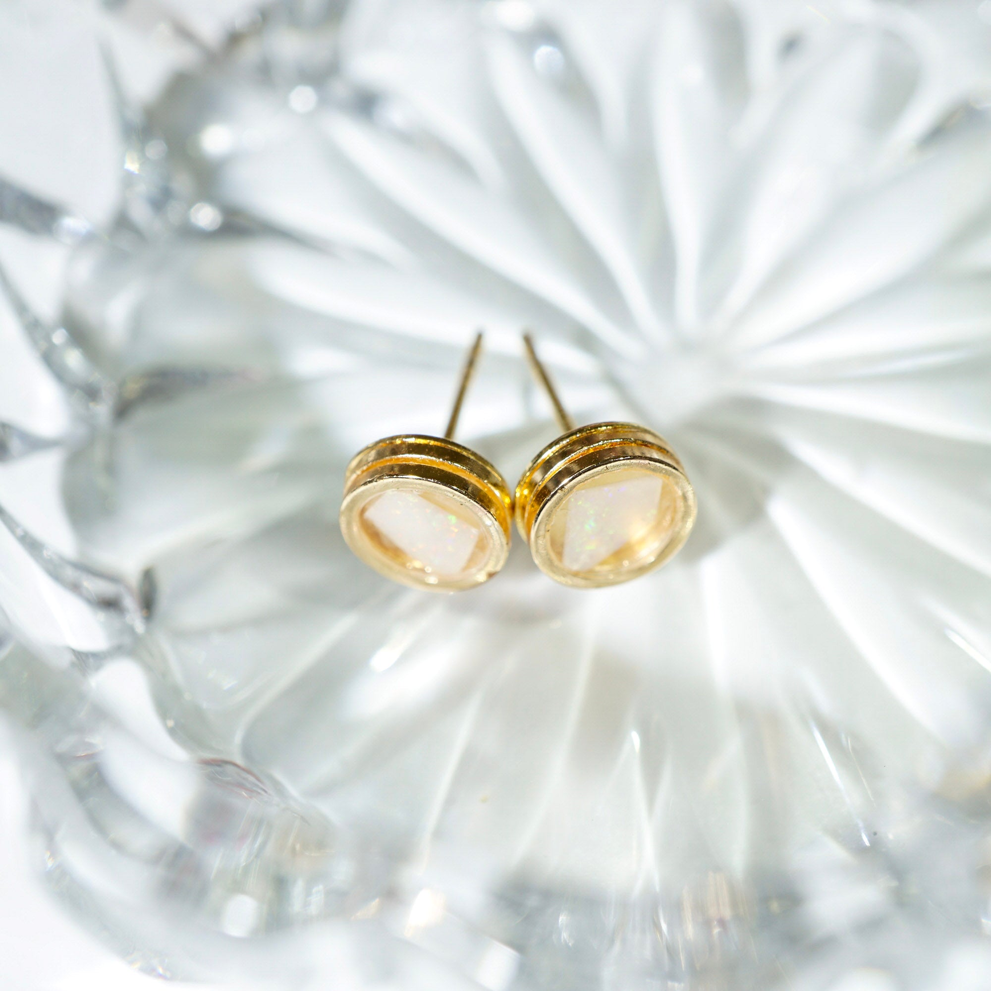 A pair of gold post earrings with oval milky white cabochon gemstones, resting on a textured clear glass plate.