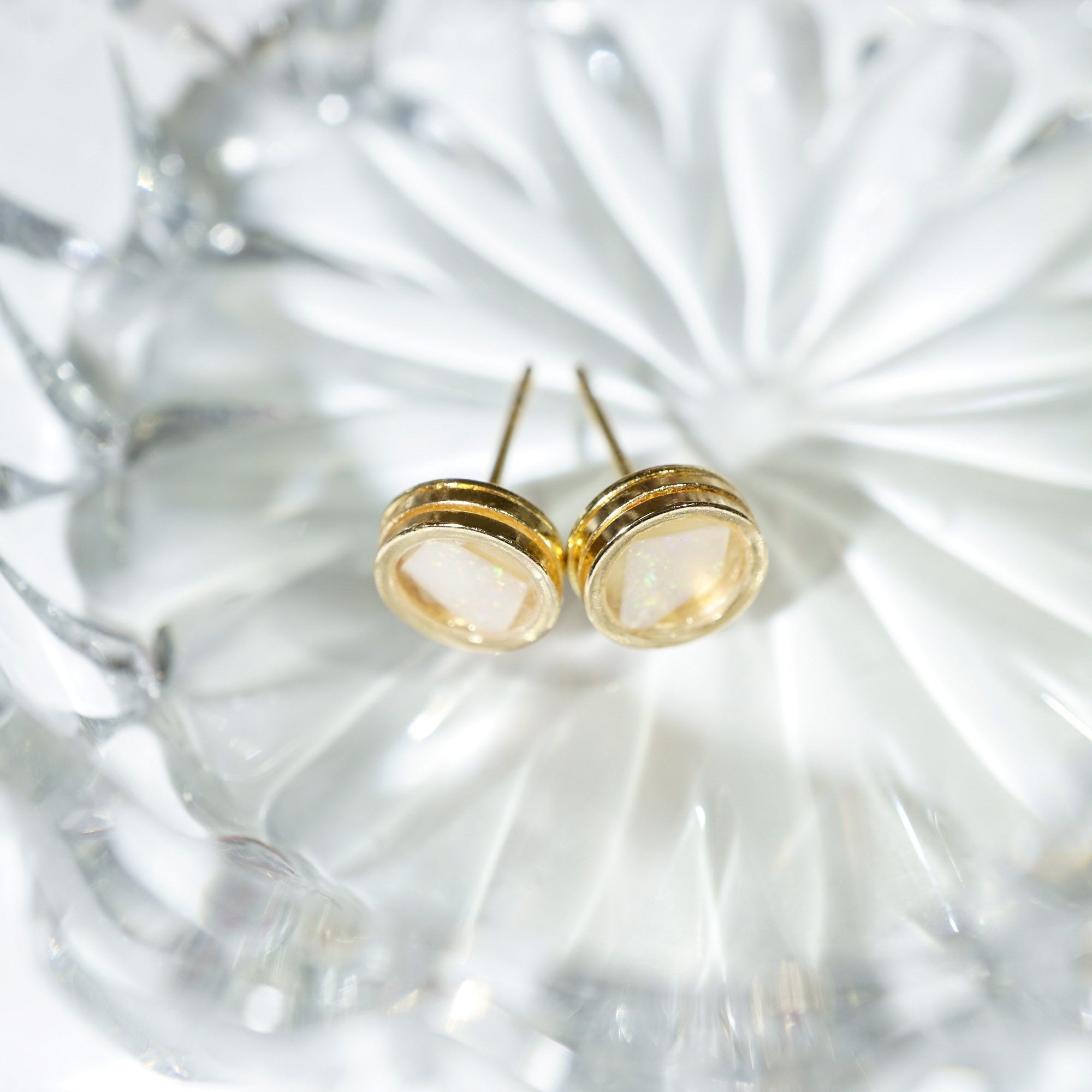 A pair of gold stud earrings with oval milky white gemstone backings, resting on a textured clear glass dish.