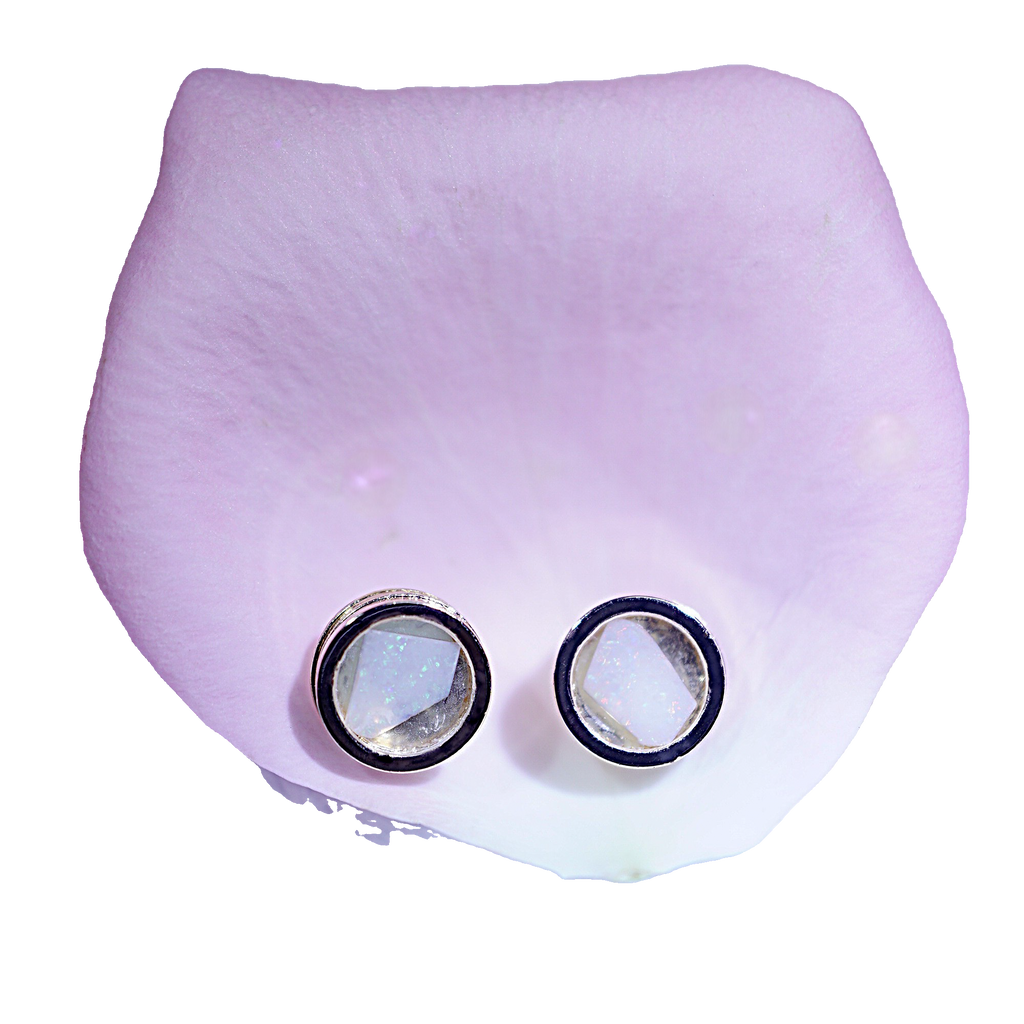A pair of round stud earrings with clear faceted stones in silver-toned bezel settings, displayed on a pale lavender petal against a white background.