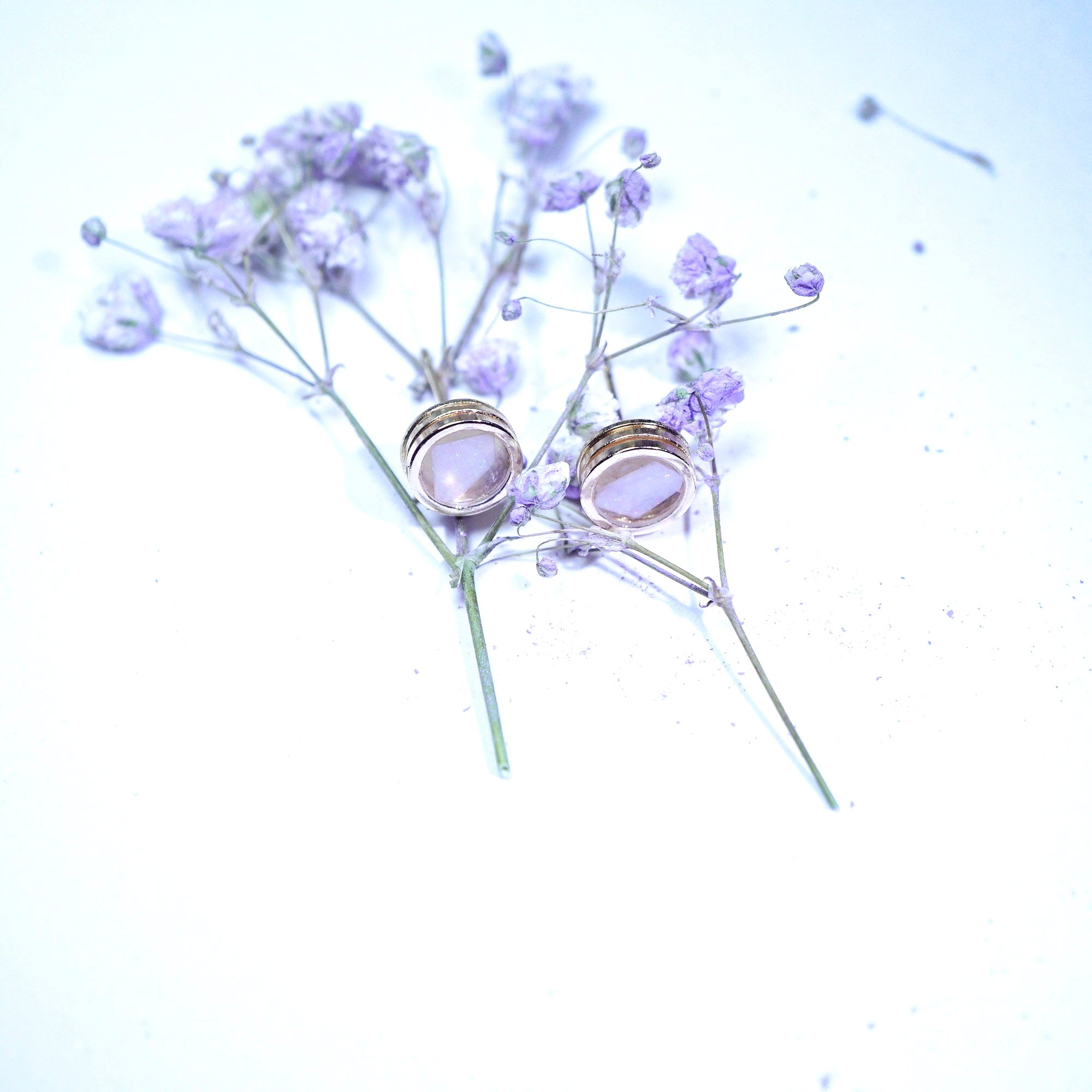 A pair of gold-toned stud earrings with oval pale lavender-purple stone inlays, displayed on delicate light purple dried flowers against a bright white background.