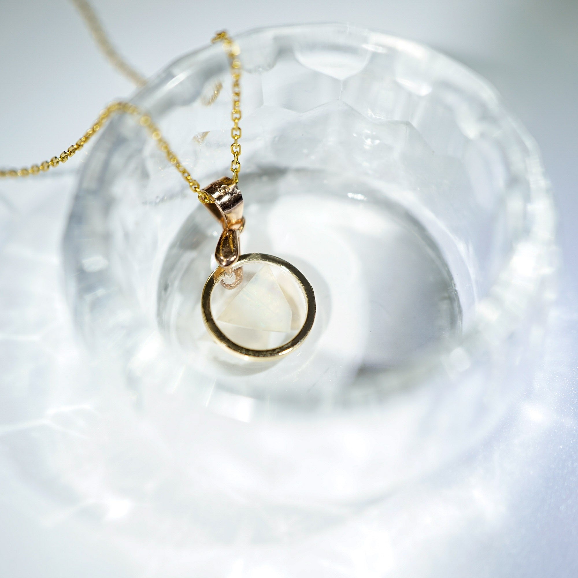 A gold necklace with a circular pendant resting inside a textured clear glass bowl on a white surface.