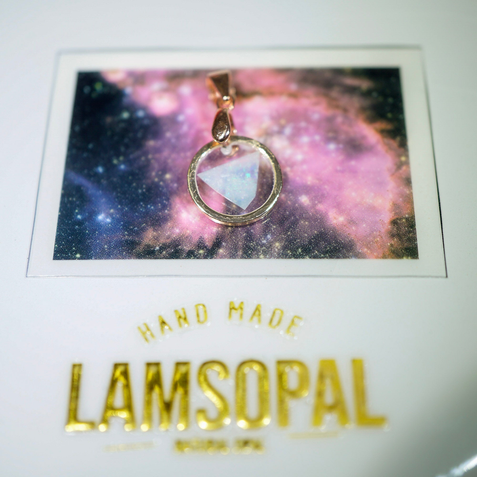 A gold-toned circular pendant with a triangular iridescent stone, displayed on a white card above a colorful galaxy print and the word 'LAMSO PAL' in gold lettering.