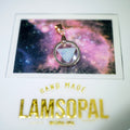 A handmade Lamsopal pendant featuring a silver-toned circular frame and a central triangular blue opal gemstone, displayed on a white card with a colorful galaxy background and gold 'HAND MADE LAMSOPAL' lettering.