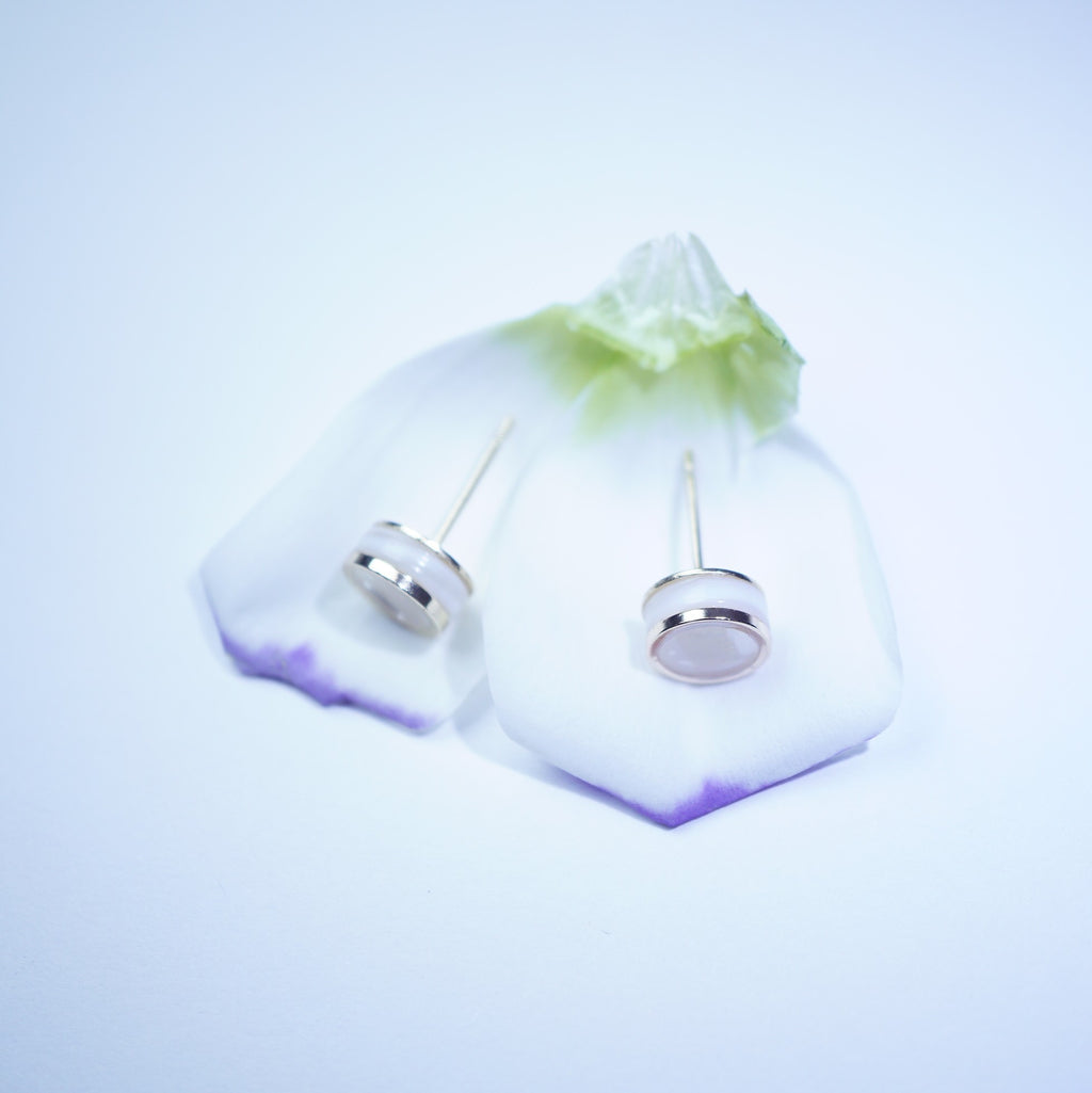 A pair of silver-toned stud earrings with oval clear stones, displayed on a translucent white stand with a green and purple gradient base against a light blue background.