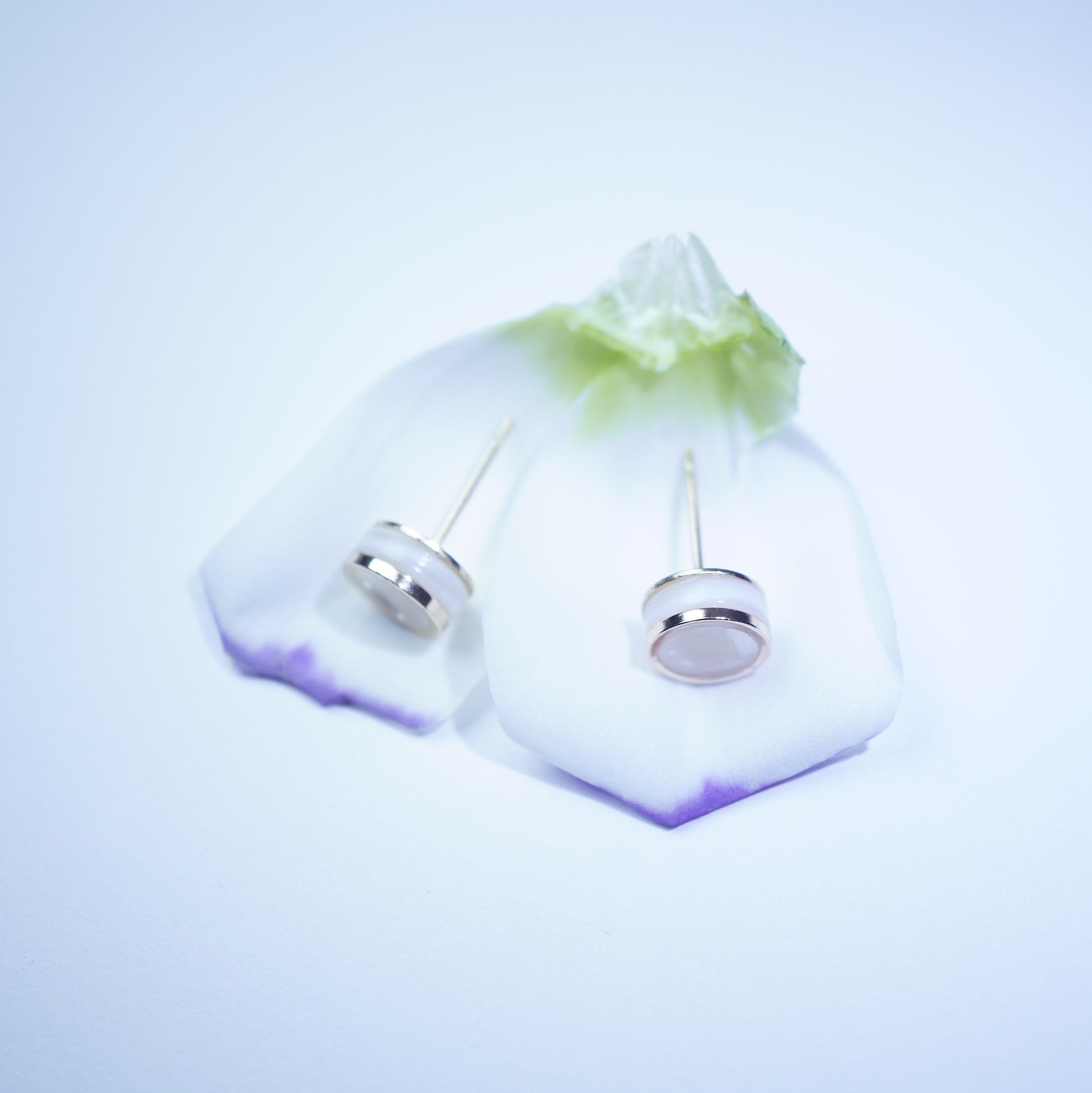 A pair of silver-toned stud earrings with oval clear stones, displayed on a translucent white stand with a green and purple gradient base against a light blue background.