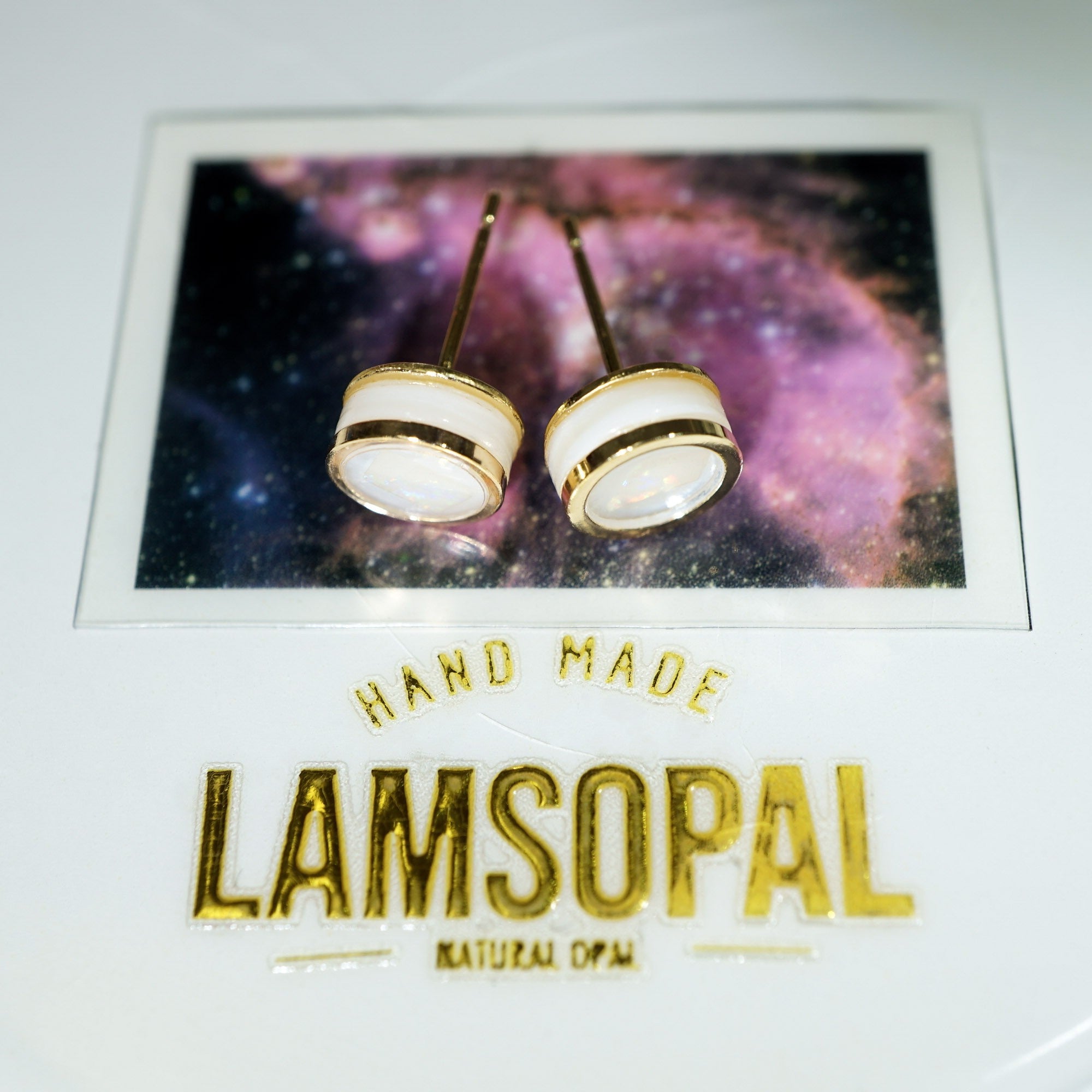 A pair of handmade stud earrings with white circular tops and gold borders, displayed on a branded card that reads 'HAND MADE LAMSOPAL NATURAL OPAL'.