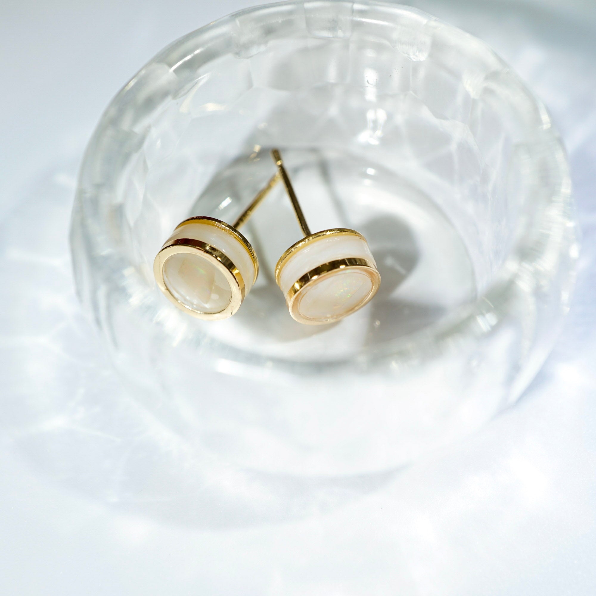 A pair of gold stud earrings with oval cream-colored stone inlays, displayed inside a clear textured glass bowl on a white surface.
