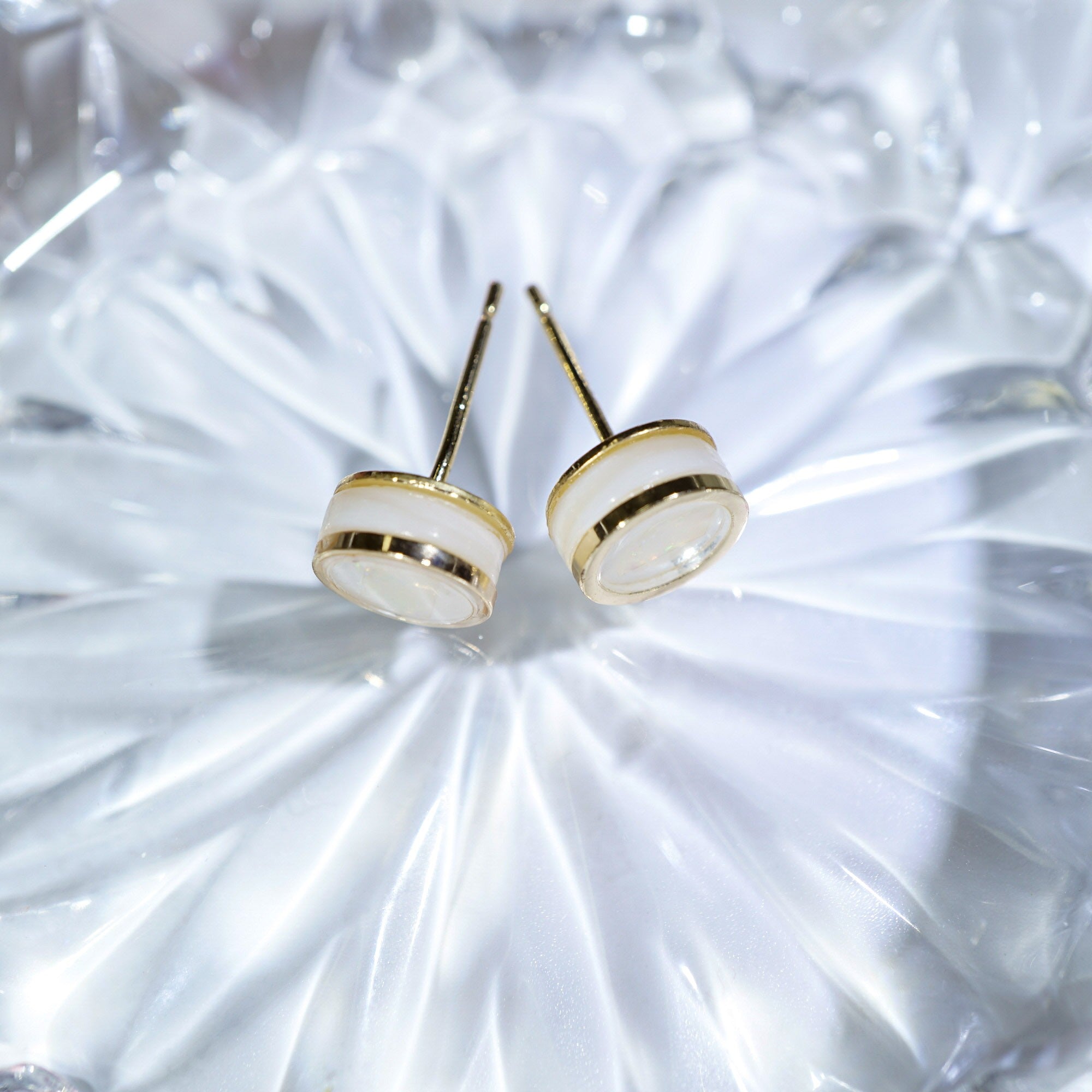 A pair of gold stud earrings with oval white inlay details, displayed on a textured, reflective white surface.