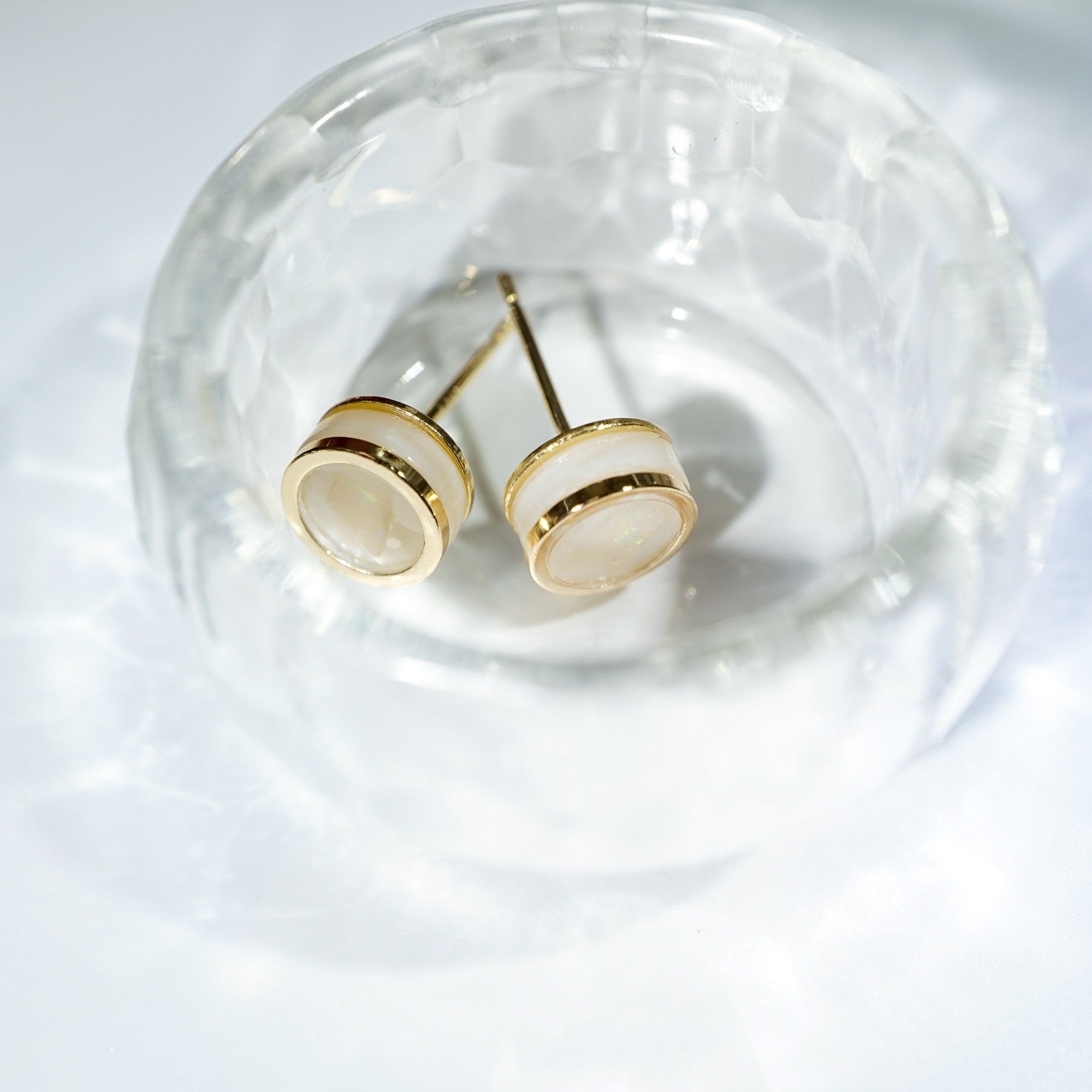 A pair of gold stud earrings with oval cream-colored stone inlays, displayed inside a clear textured glass bowl on a white surface.