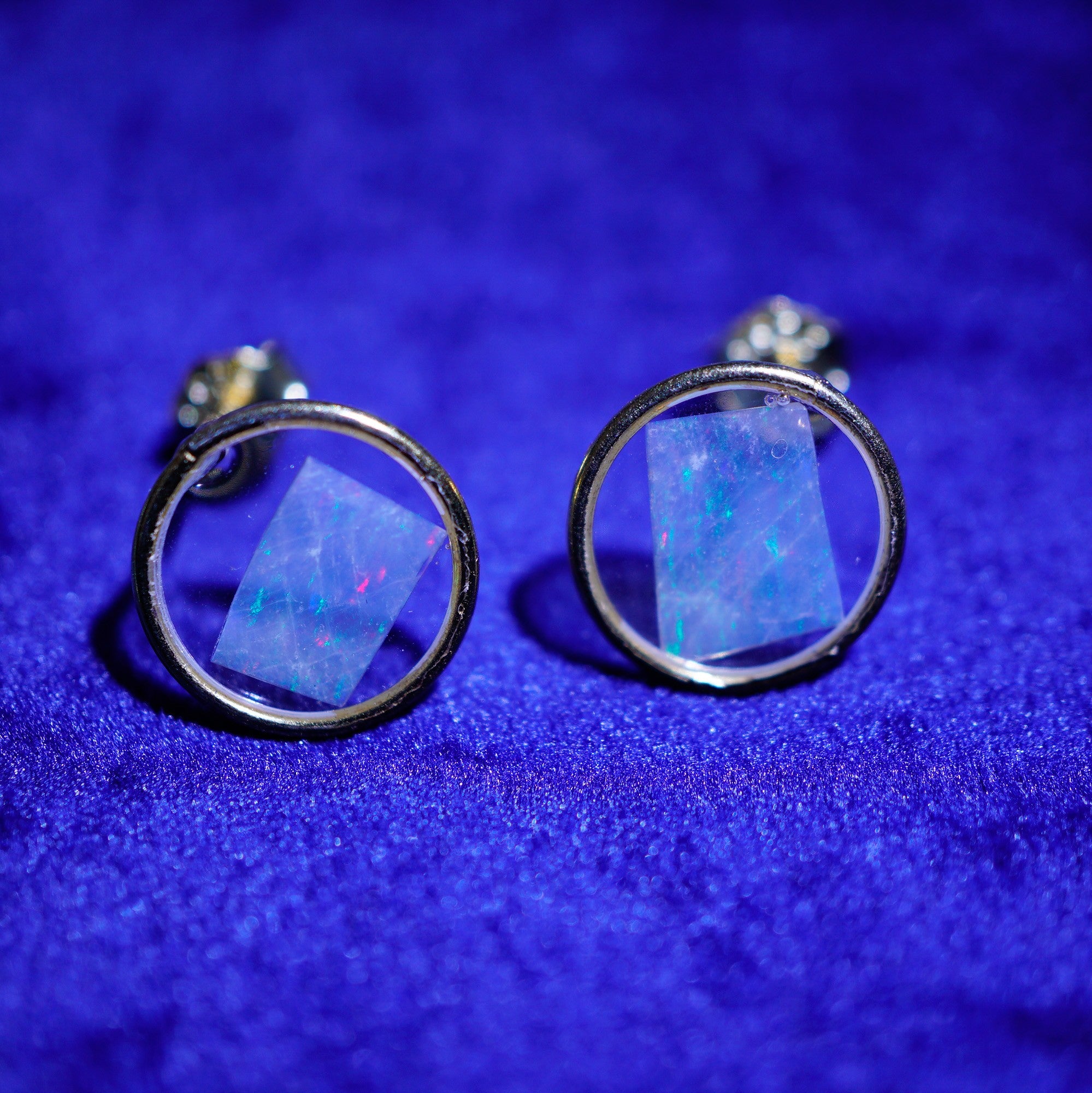A pair of silver-toned circular stud earrings with faceted blue iridescent opal-like centers, displayed on a textured royal blue velvet surface.