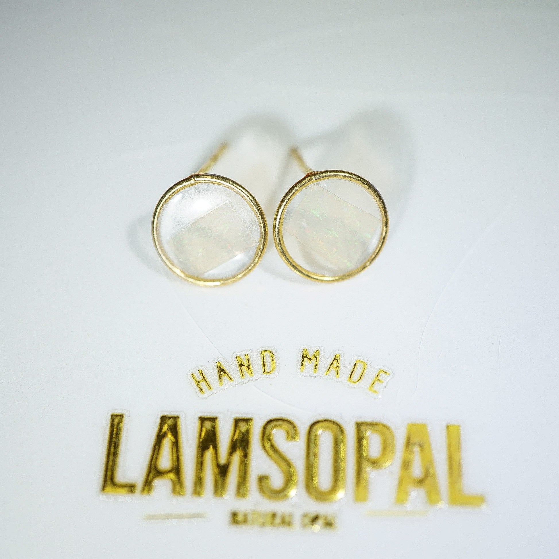 A pair of gold round stud earrings with milky white stone centers, displayed on a white surface above gold foil text that reads 'HAND MADE LAMSOPAL'.