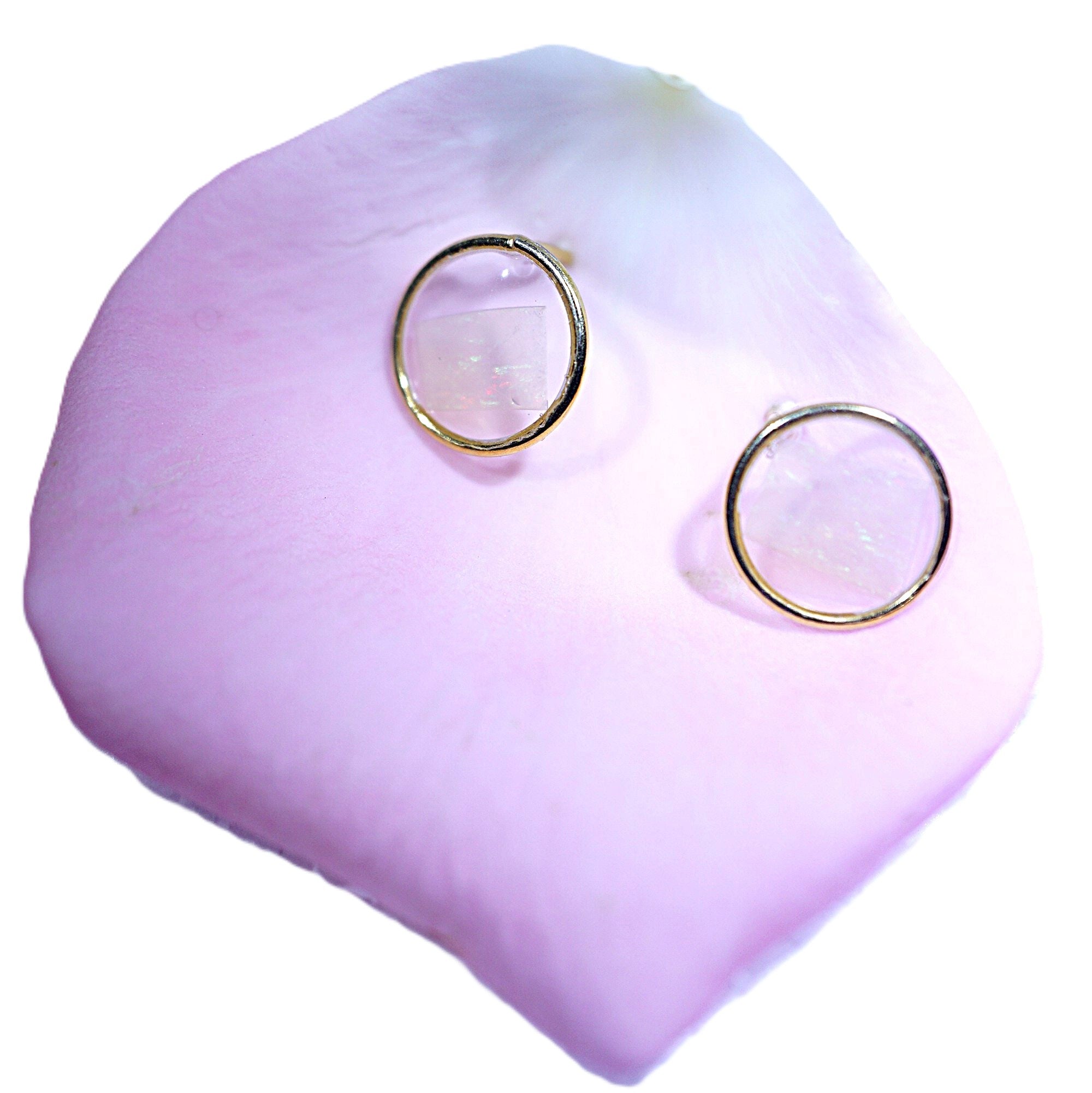 A pair of gold hoop earrings with clear faceted gemstone centers, displayed on a soft pink rose petal against a white background.