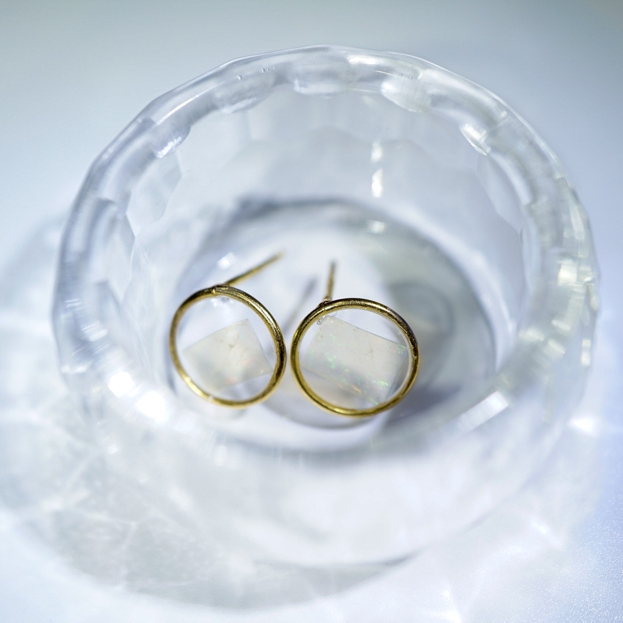 A pair of gold hoop earrings with milky white oval inlays, displayed inside a clear faceted glass bowl on a white surface.